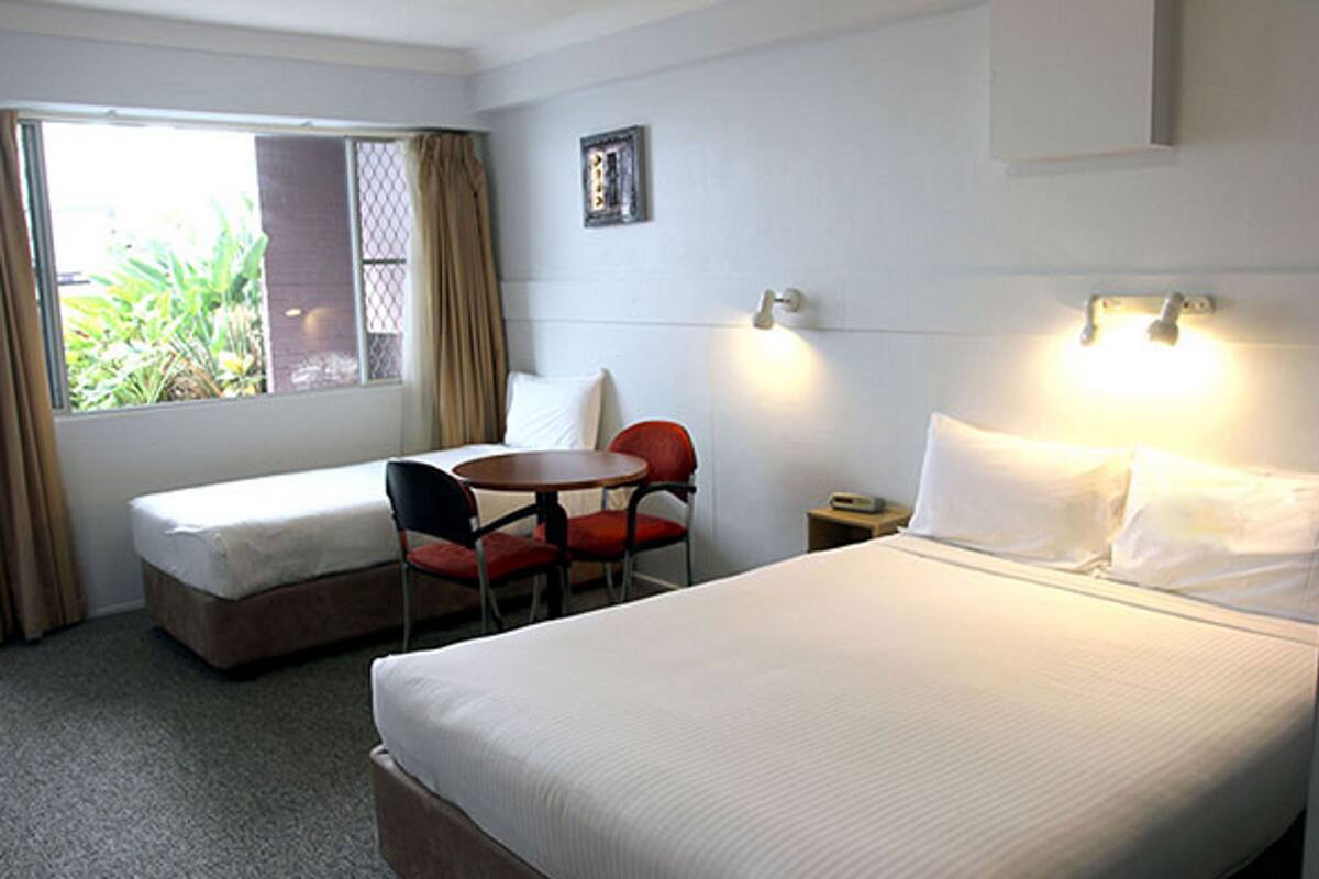 A hotel room with a single bed, a small desk, a chair, a window with curtains, and a bedside table.
