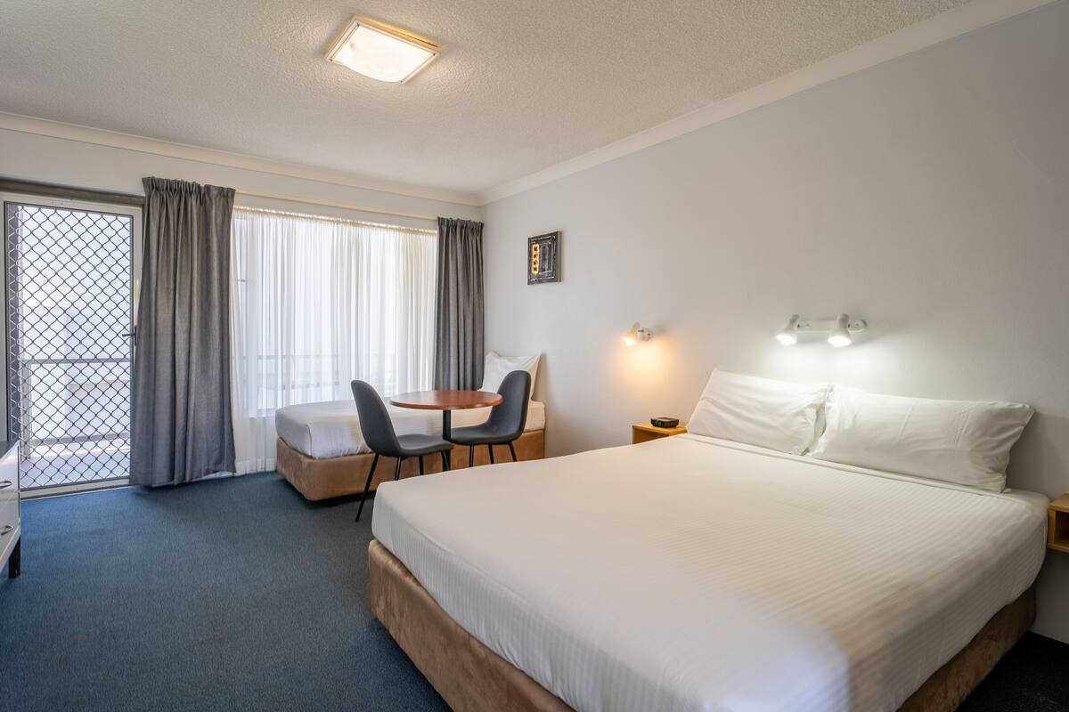 A hotel room with a large bed, a desk with two chairs, a small table, and a window with curtains.