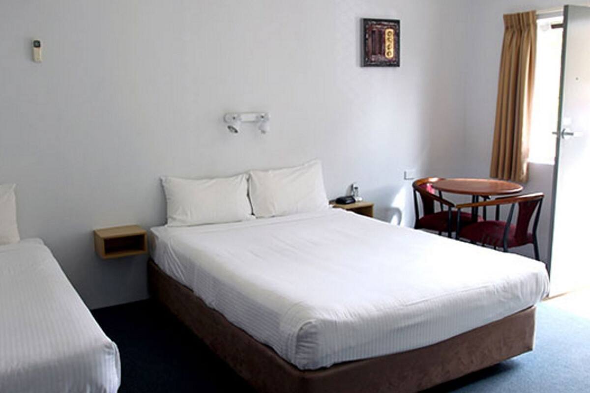 A hotel room with two single beds, a small table, a chair, and a wall-mounted light fixture.