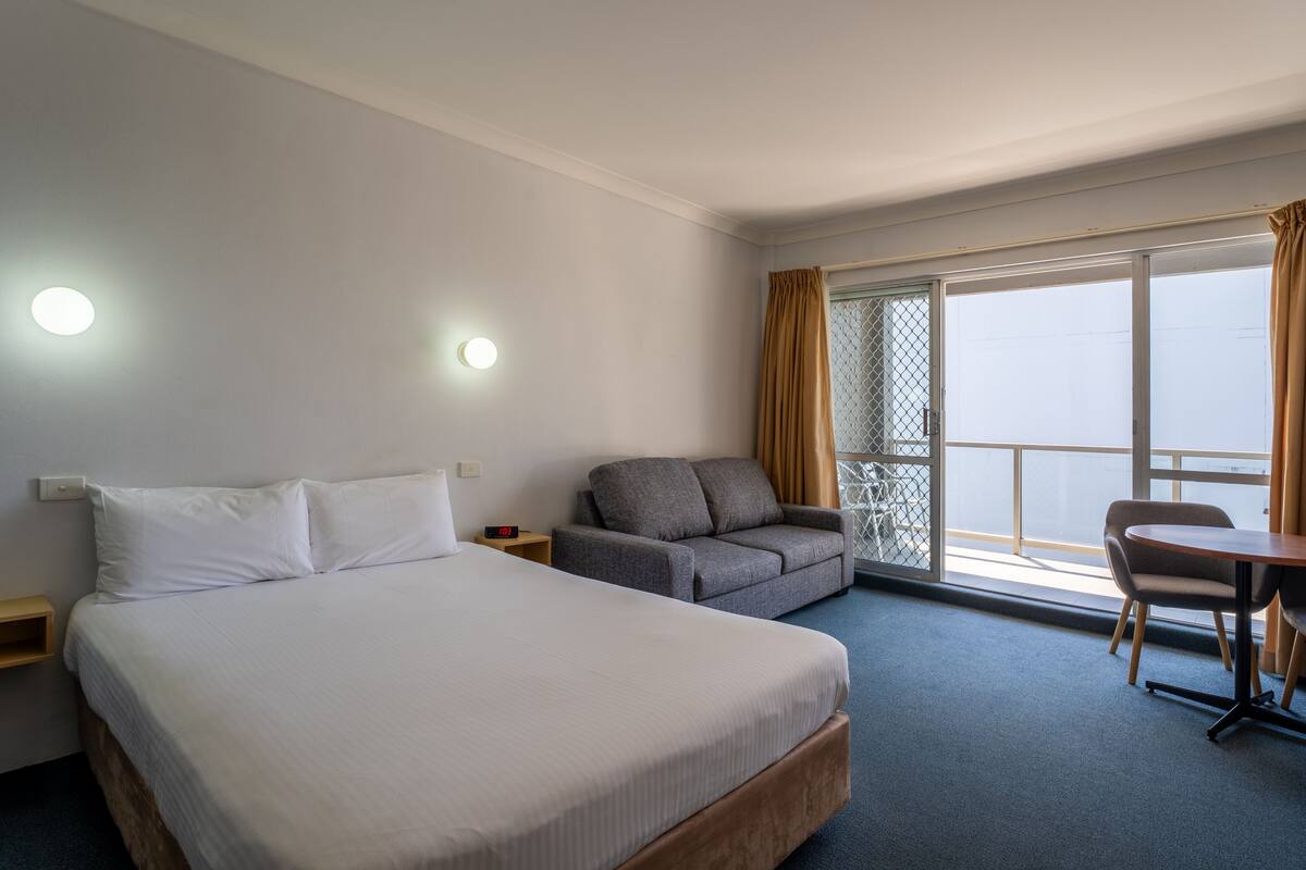A hotel room with a large bed, a gray sofa, a small table, and a balcony with a view.