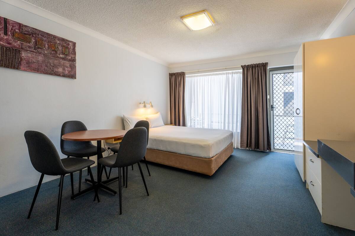 A compact hotel room with a single bed, a small desk with a chair, a table with four chairs, and a window with curtains.