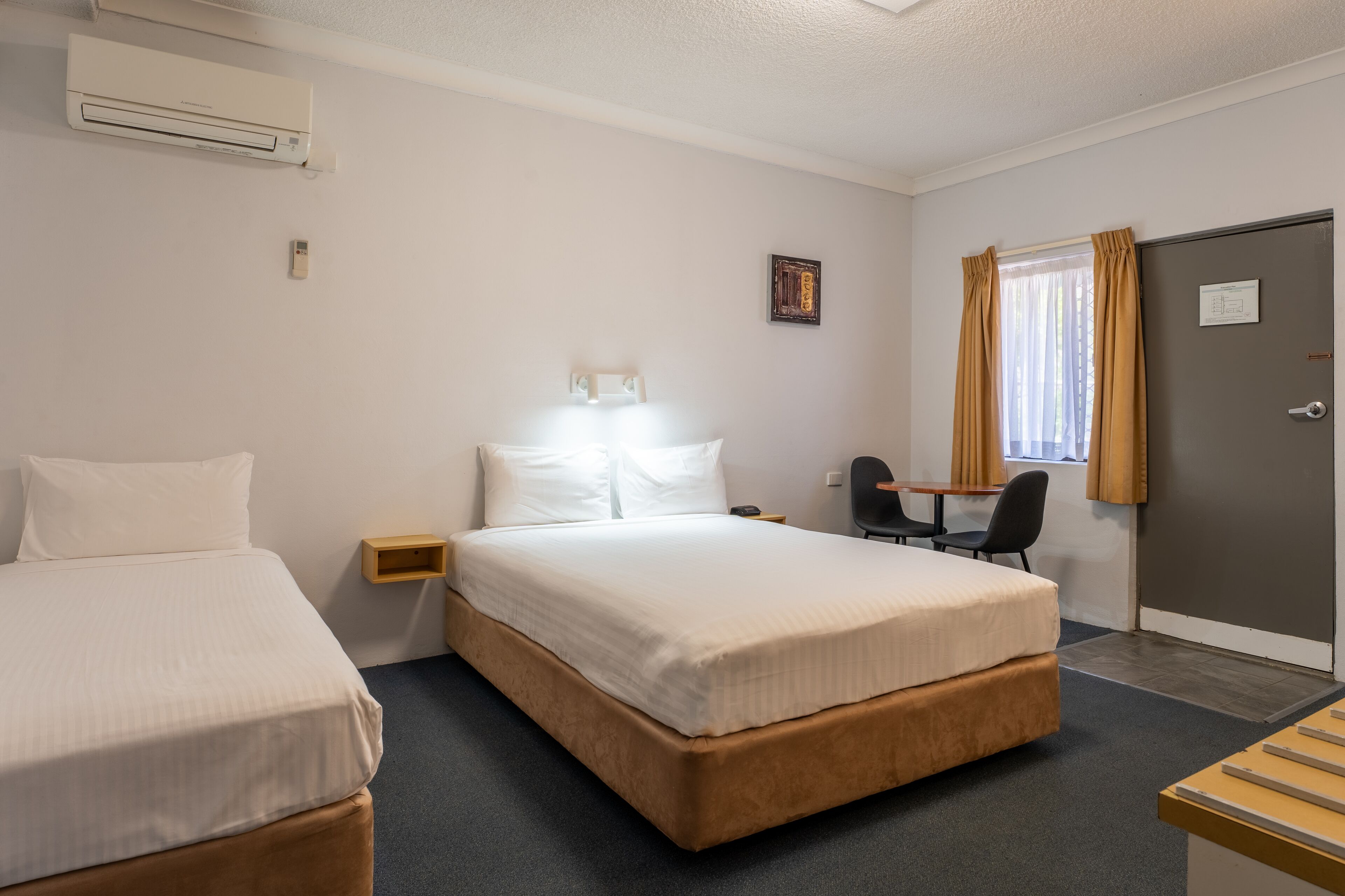 Hotellihuone, jossa on kaksi erillistä sänkyä, työpöytä tuolilla, pieni pöytä, ikkuna verhoilla ja ilmastointilaite.