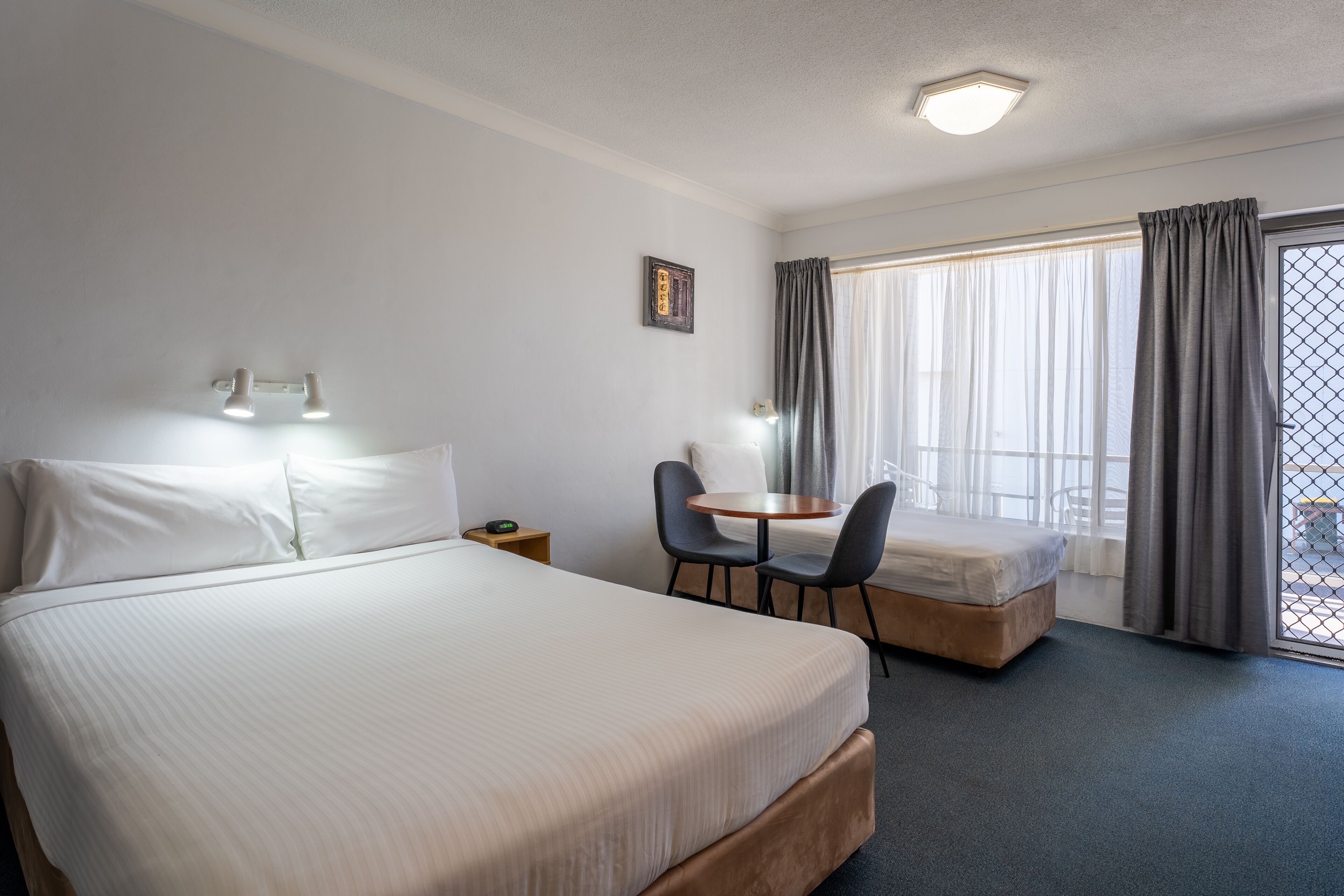 Habitación de hotel con una cama grande, una cama más pequeña, un escritorio con dos sillas, una ventana con cortinas y una mesita.