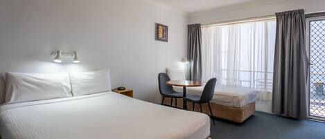 Habitación de hotel con una cama grande, una cama más pequeña, un escritorio con dos sillas, una ventana con cortinas y una mesita.