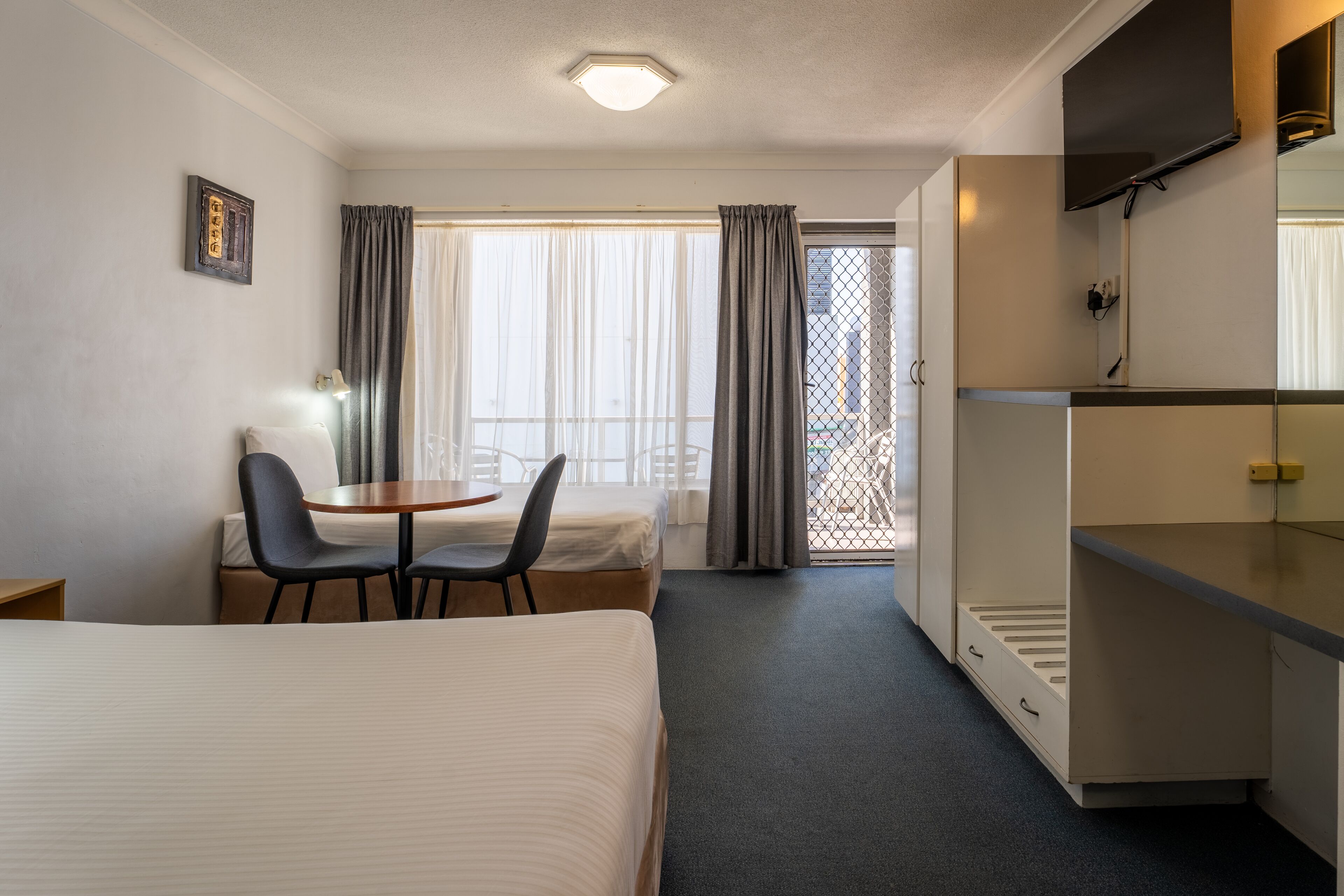 A hotel room with a bed, a desk, two chairs, a small table, a TV, and a balcony with curtains.