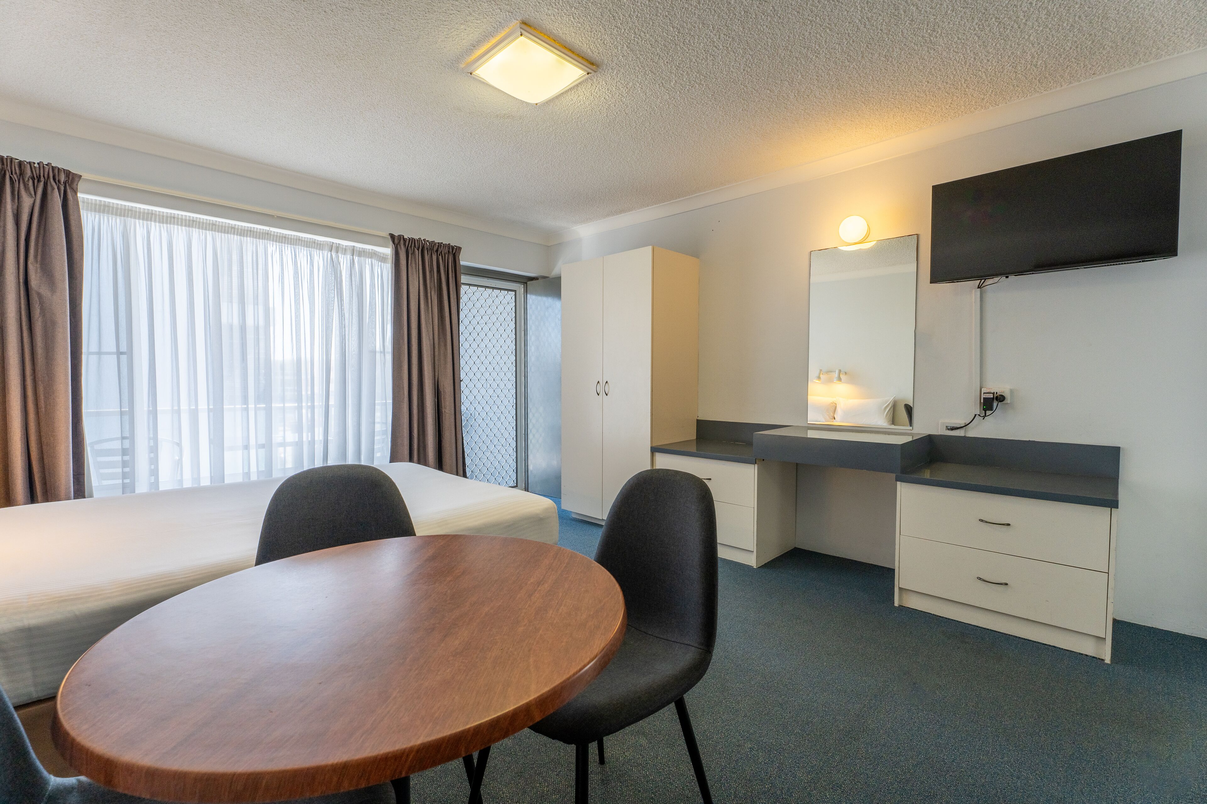 A hotel room with a bed, a desk, a chair, a table, a TV, and a window with curtains.