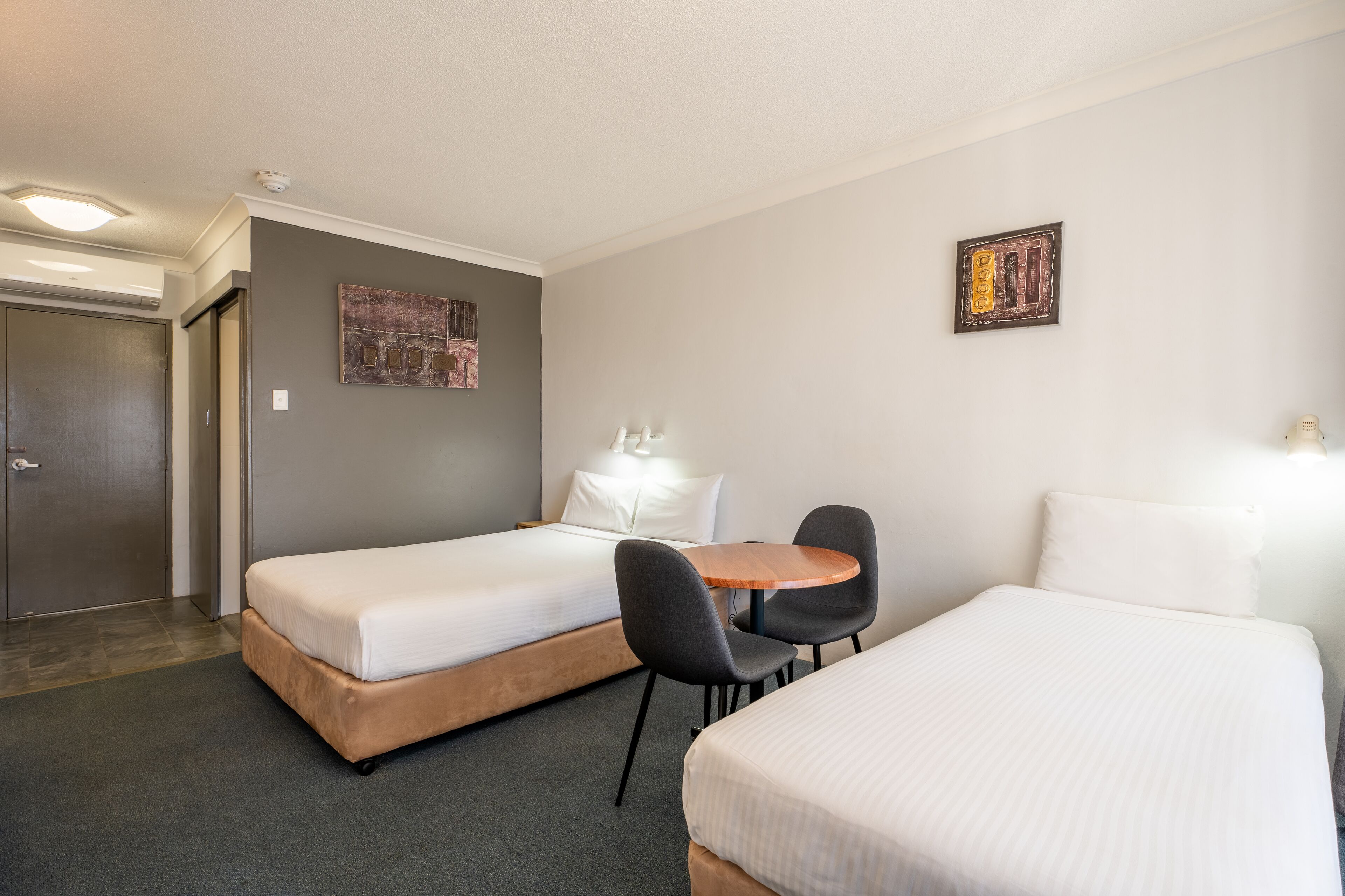 A hotel room with two beds, a small table, chairs, a door, and a wall-mounted artwork.
