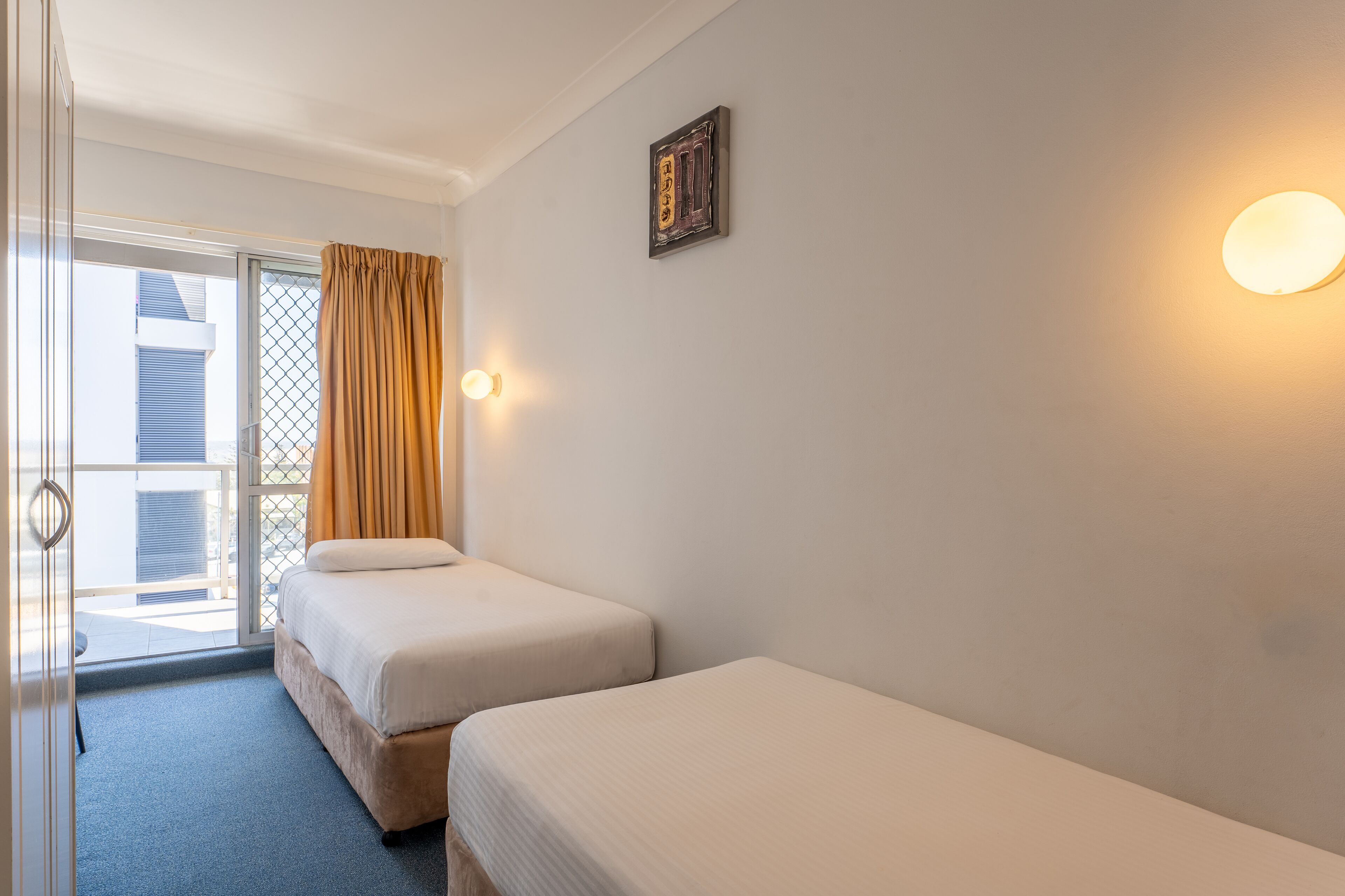 A hotel room with two beds, a sliding glass door to the balcony, and a wall-mounted light fixture.