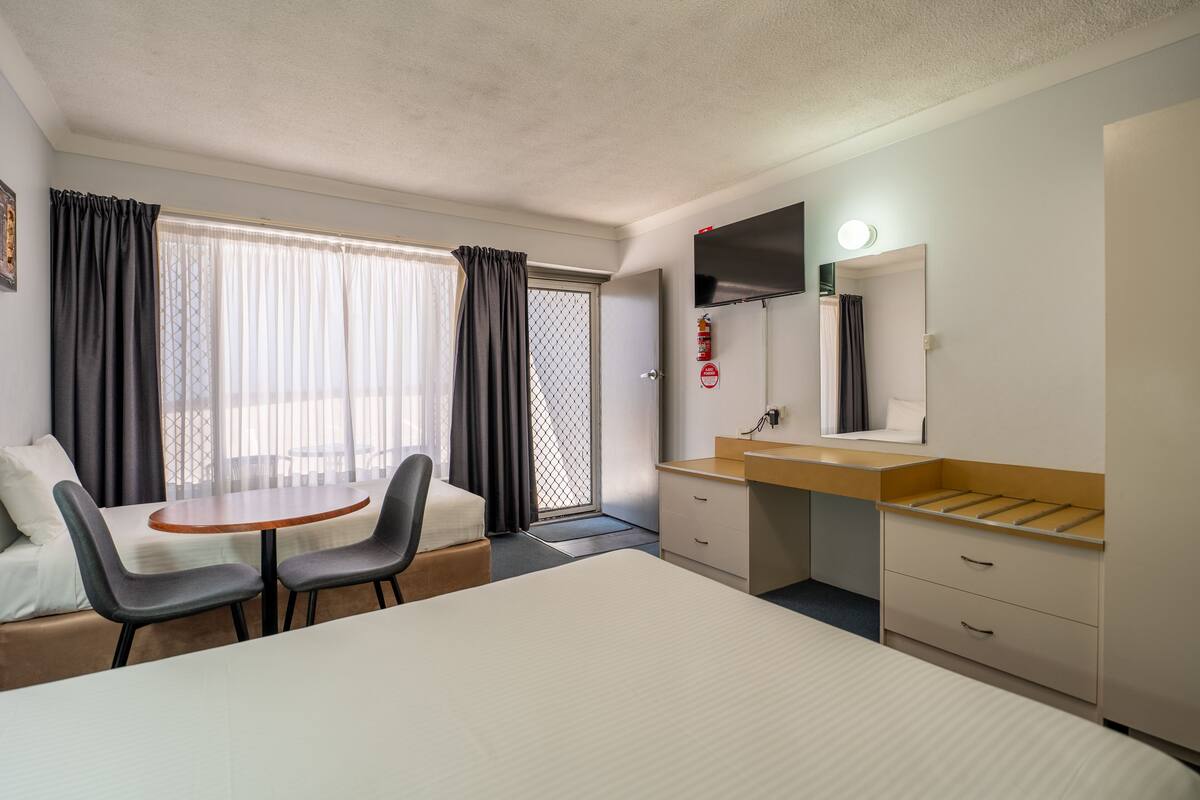 A hotel room with a bed, a desk, two chairs, a small table, a bathroom with a sink and mirror, and a window with curtains.