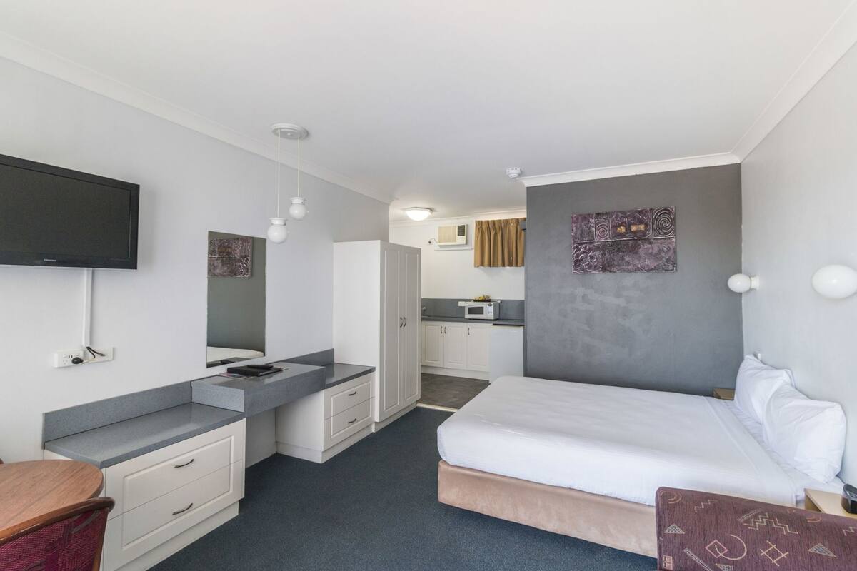 A hotel room with a bed, a desk, a chair, a TV, a microwave, and a kitchenette.