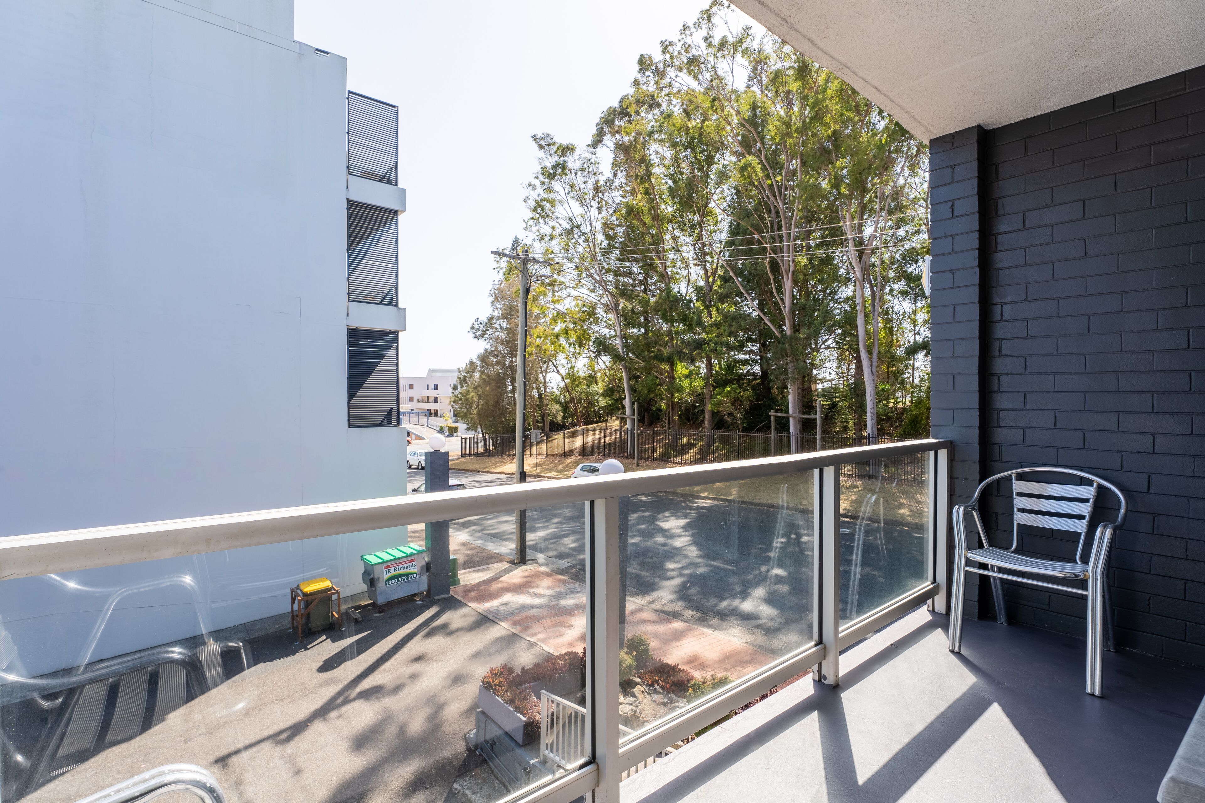 A balcony with a glass railing, a metal bench, and a view of a street with trees and buildings.