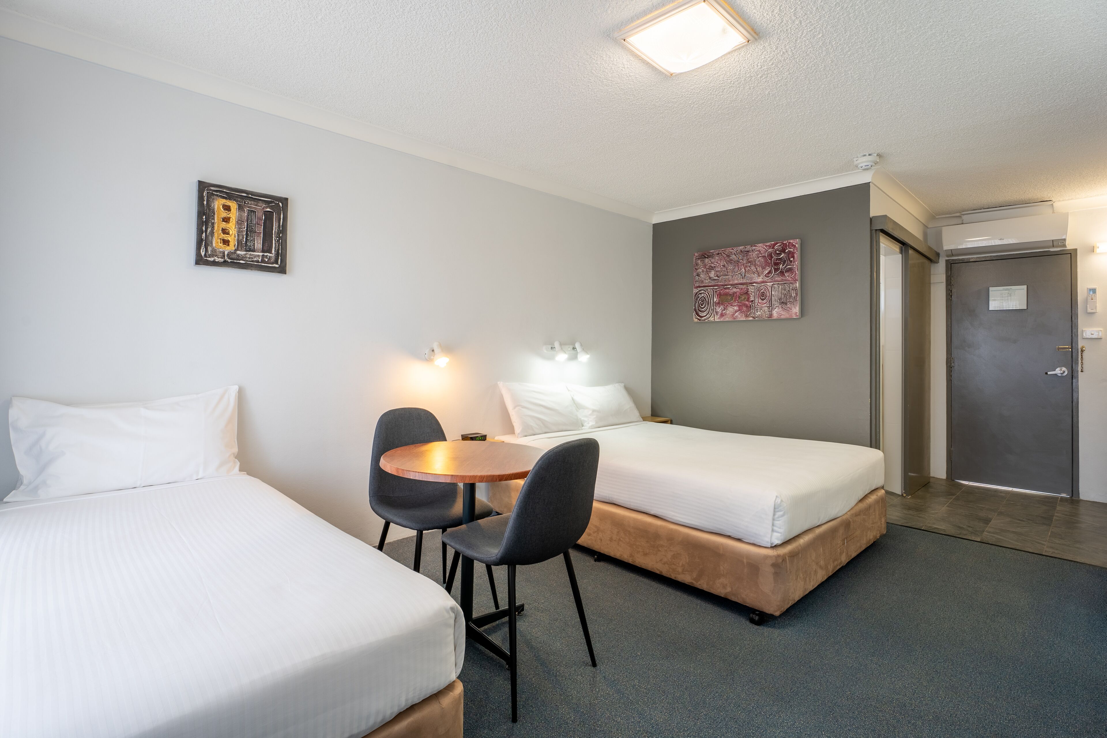 Ein Hotelzimmer mit zwei Einzelbetten, einem kleinen runden Tisch mit Stühlen, einer Wandleuchte und einer Tür mit einem Schild.