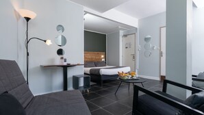 A modern hotel room with a grey sofa, a bed with a wooden headboard, a small table with breakfast, and a floor lamp. - Celenya Hôtel (Toulon)