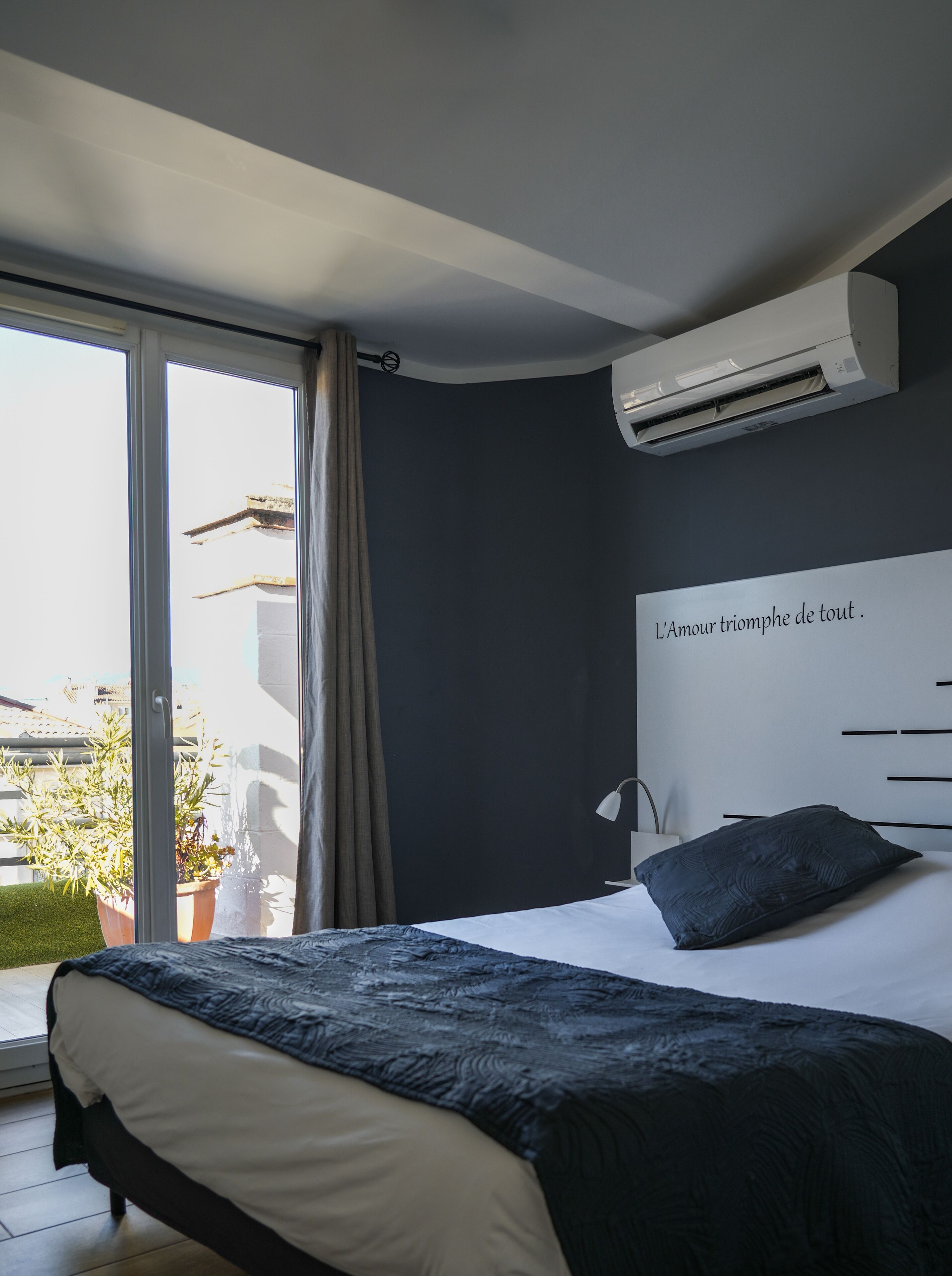 Ein Schlafzimmer mit einem Bett, einer Wand-Klimaanlage, einem Fenster mit Vorhängen und Blick auf Gebäude.