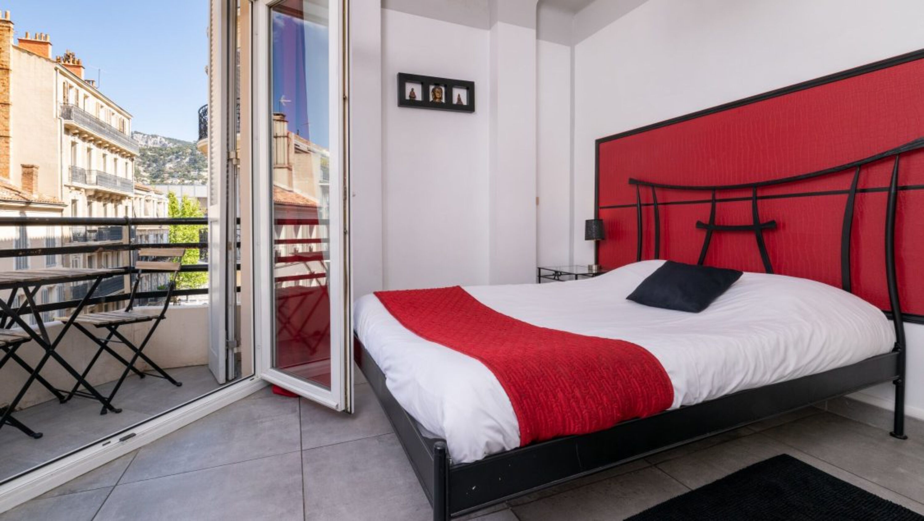 A bedroom with a bed, a red headboard, a balcony with a table and chairs, and a city view.