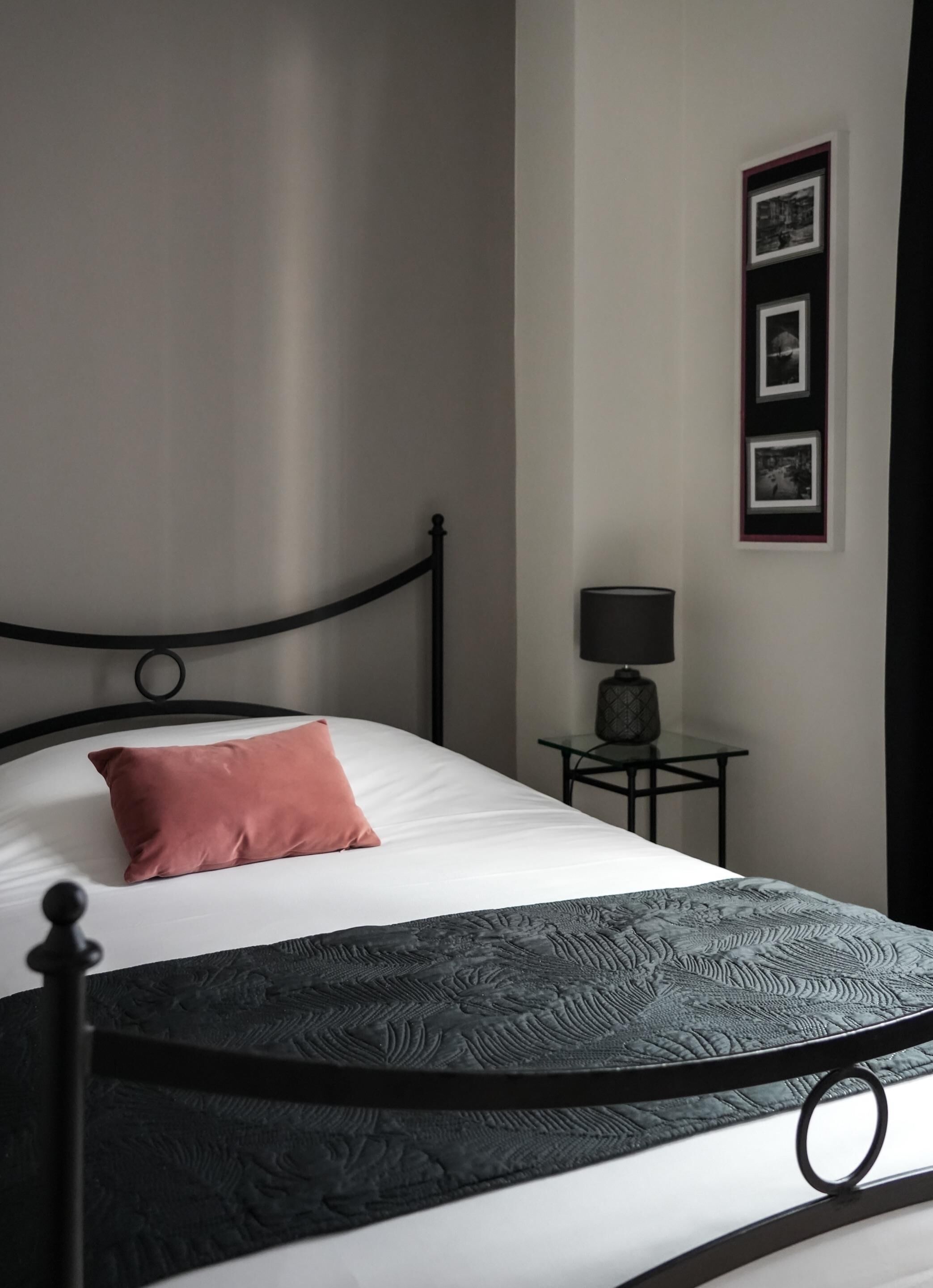 Una cama bien tendida con estructura de metal negro, colchón blanco y almohada rosa. Junto a la cama, una mesita con una lámpara.