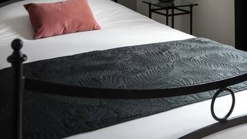 A neatly made bed with a black metal frame, a white mattress, and a pink pillow. There is a small table with a lamp beside the bed.
