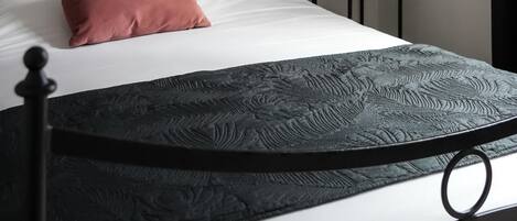 Una cama bien tendida con estructura de metal negro, colchón blanco y almohada rosa. Junto a la cama, una mesita con una lámpara.