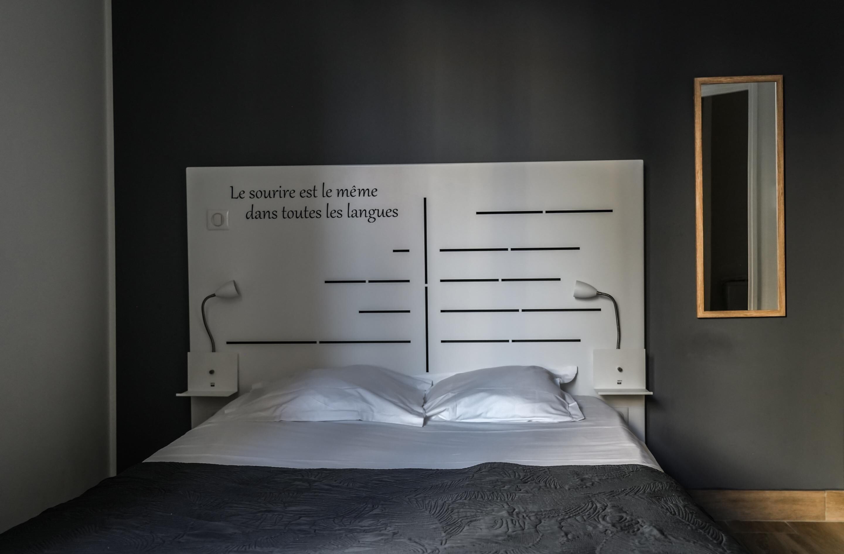 Une chambre moderne avec un grand lit, une tête de lit ornée de motifs et d’inscriptions, une table de chevet et un miroir au mur.