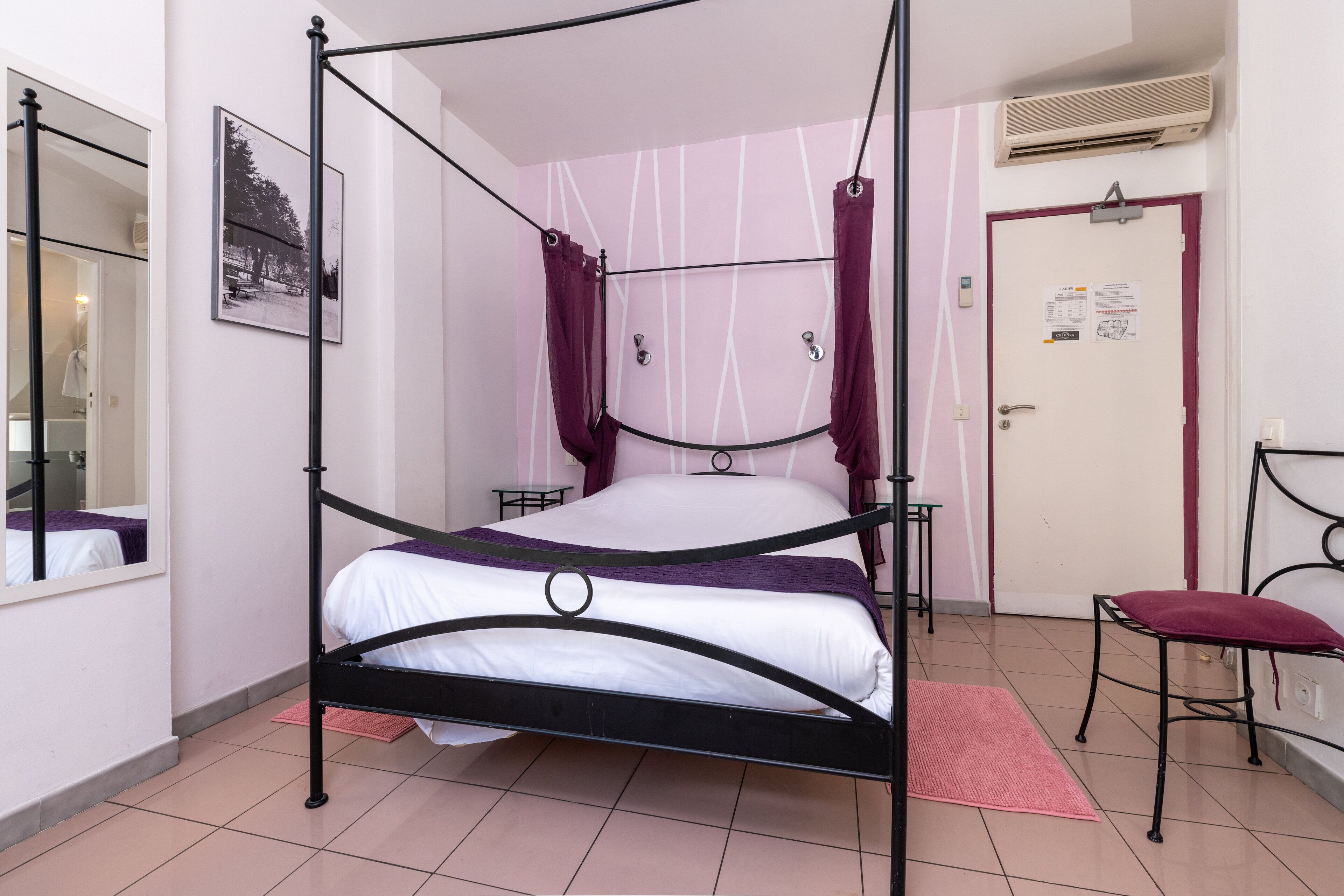 Un llit amb cortines a quatre pals, matalàs blanc i roigda morada, una petita taula de nit, un mirall i una porta amb un tabló d'anuncis.