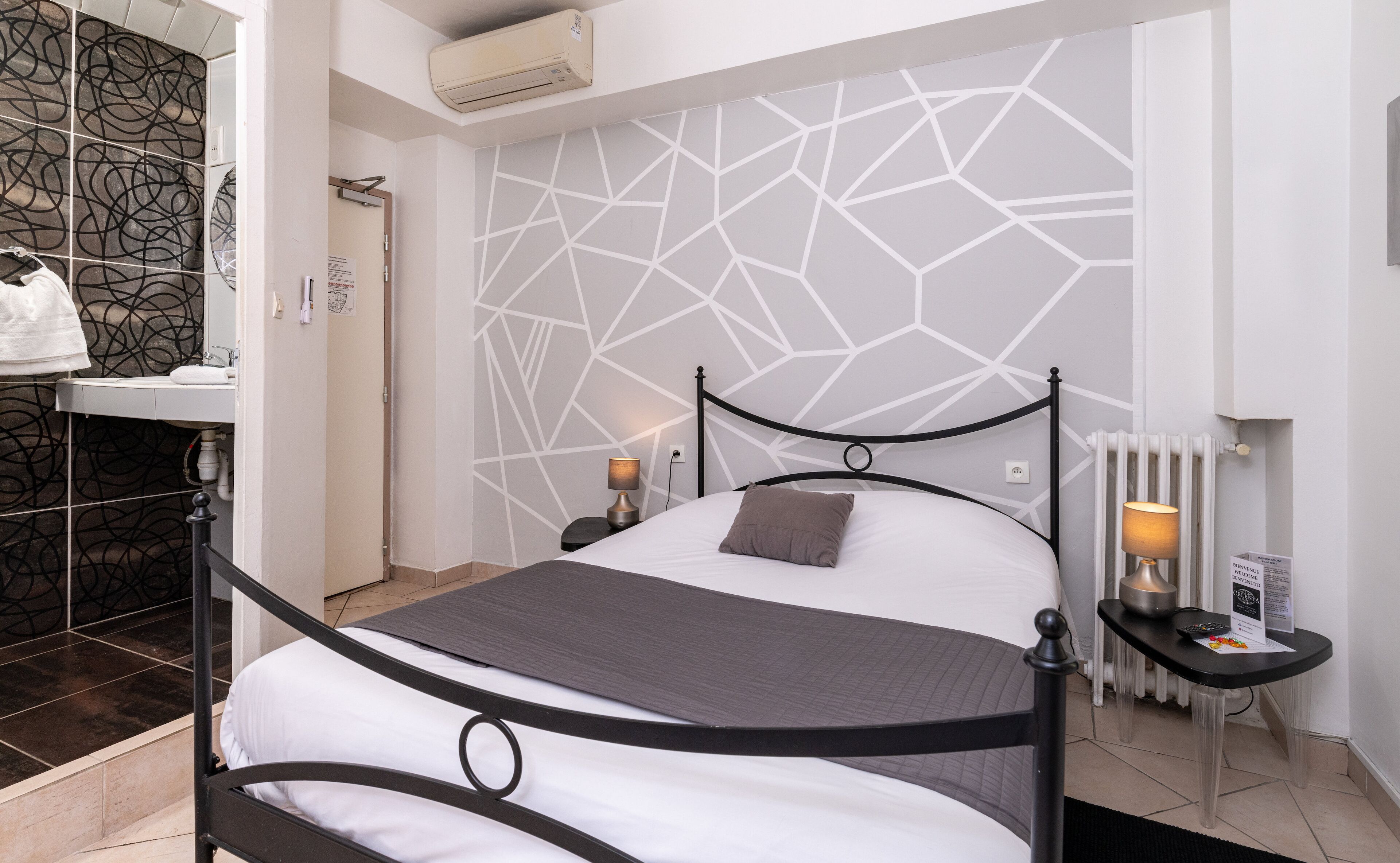 Une chambre moderne avec un mur orné d’un motif géométrique, un lit en métal noir, une table de chevet avec une lampe et un radiateur.