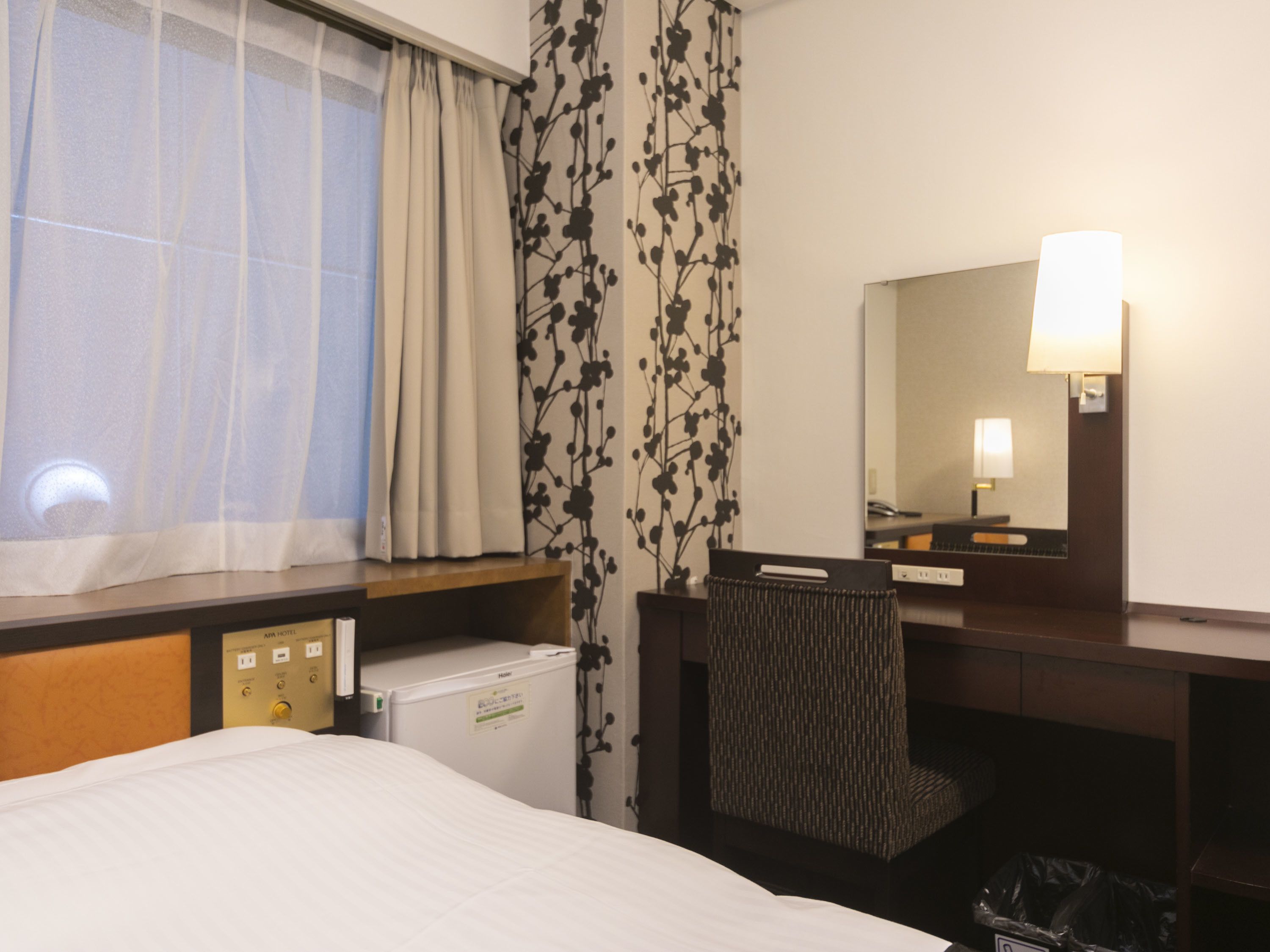 A hotel room with a bed, a desk, a chair, a mirror, a window with curtains, and a small refrigerator.