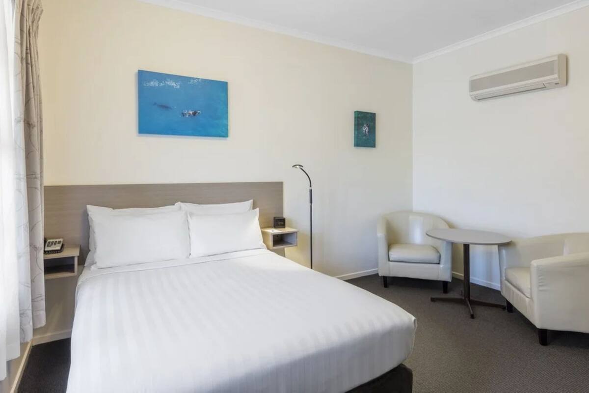 A hotel room with a large bed, two armchairs, a small table, a nightstand, and a wall-mounted air conditioner.