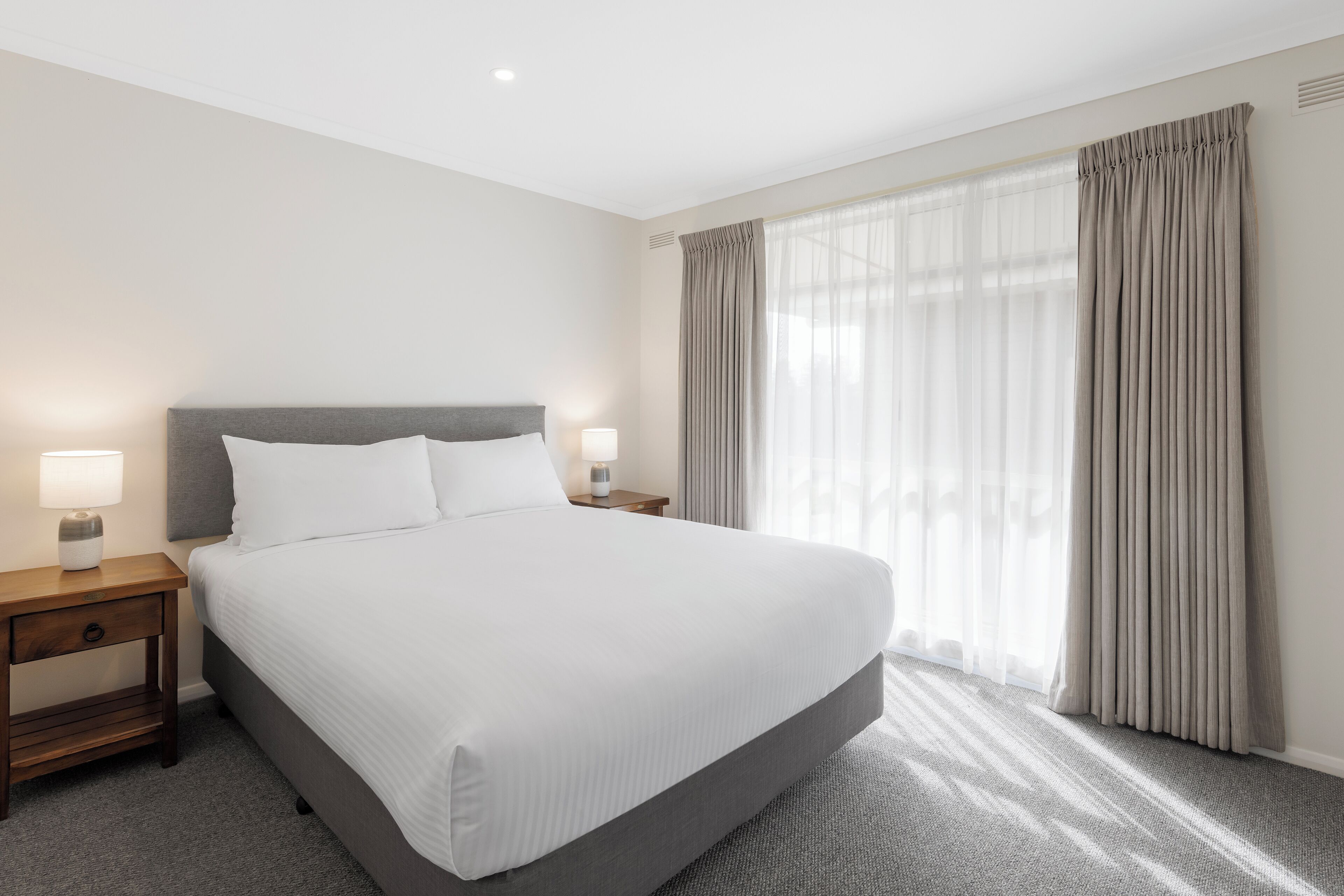 Una cama bien hecha con sábanas blancas, una mesita de noche de madera con una lámpara y un ventanal con cortinas translúcidas.