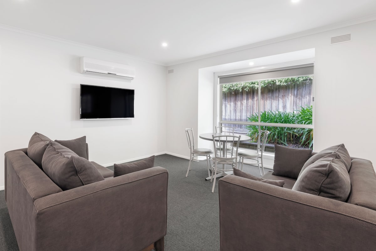 A modern living room with a grey sofa set, a flat-screen TV mounted on the wall, and a dining area with a table and chairs.