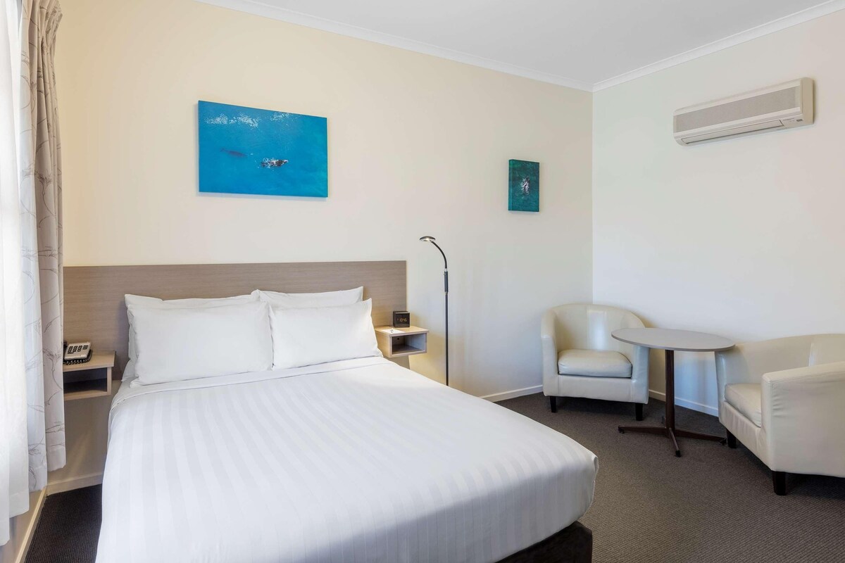A hotel room with a large bed, two chairs, a small table, a nightstand, a wall-mounted air conditioner, and two paintings.