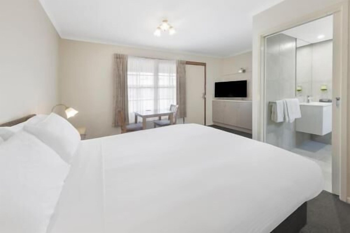 A hotel room with a large bed, a desk with a chair, a television, and a bathroom with a mirror and sink.