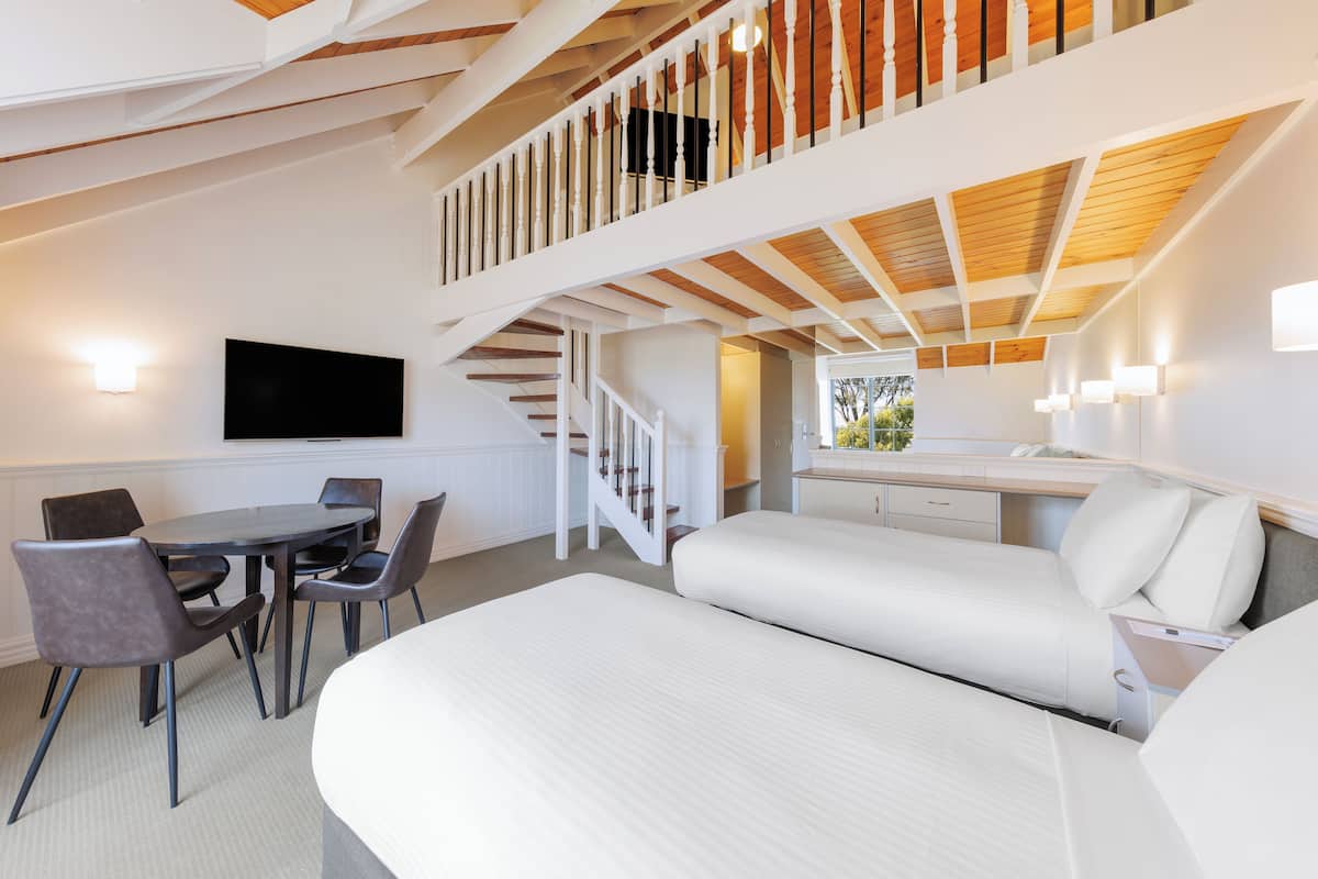 A modern hotel room with a large bed, a desk with a chair, a dining table with chairs, and a staircase leading to an upper level.