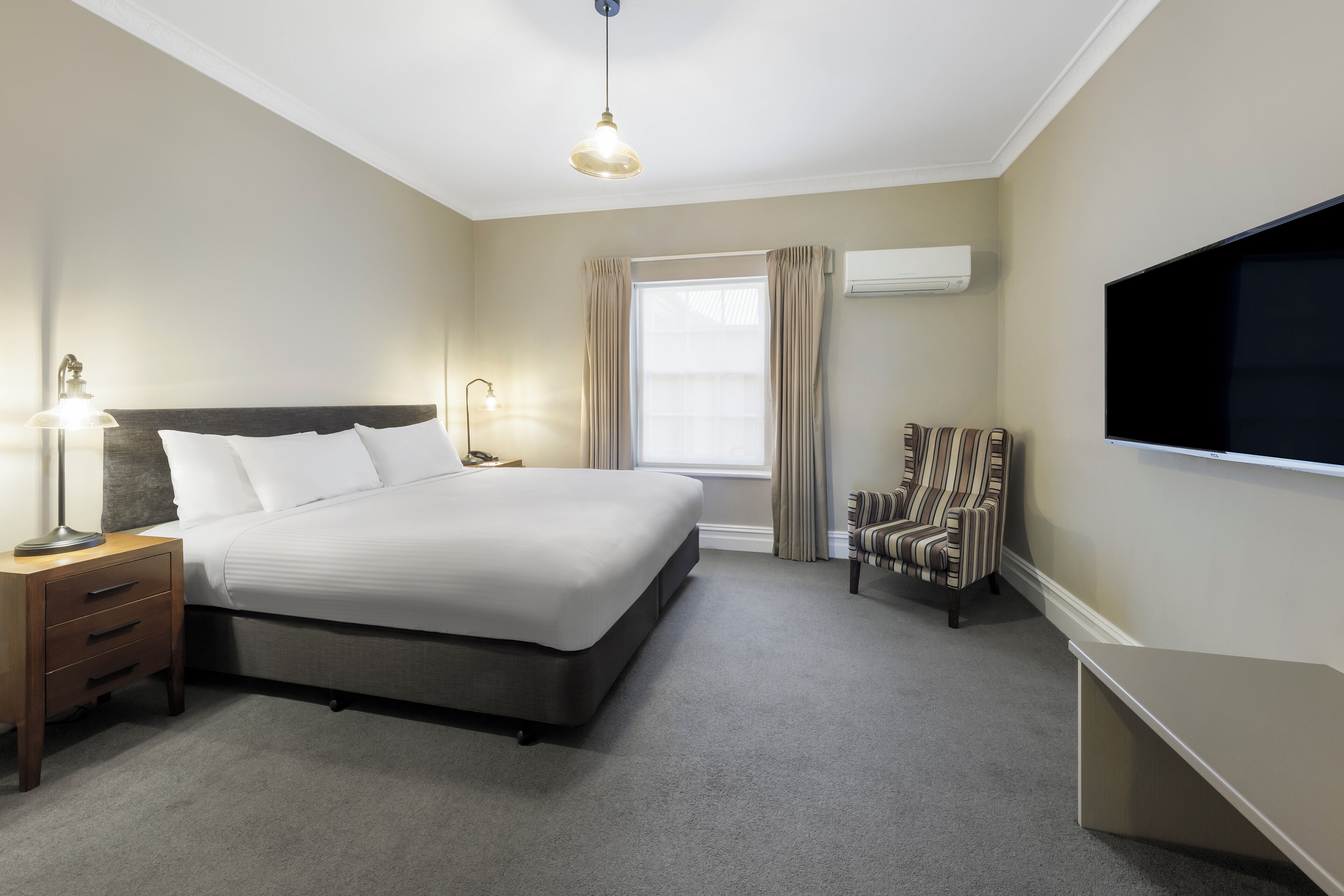 A hotel room with a large bed, bedside lamps, a chair, a TV, and an air conditioning unit.