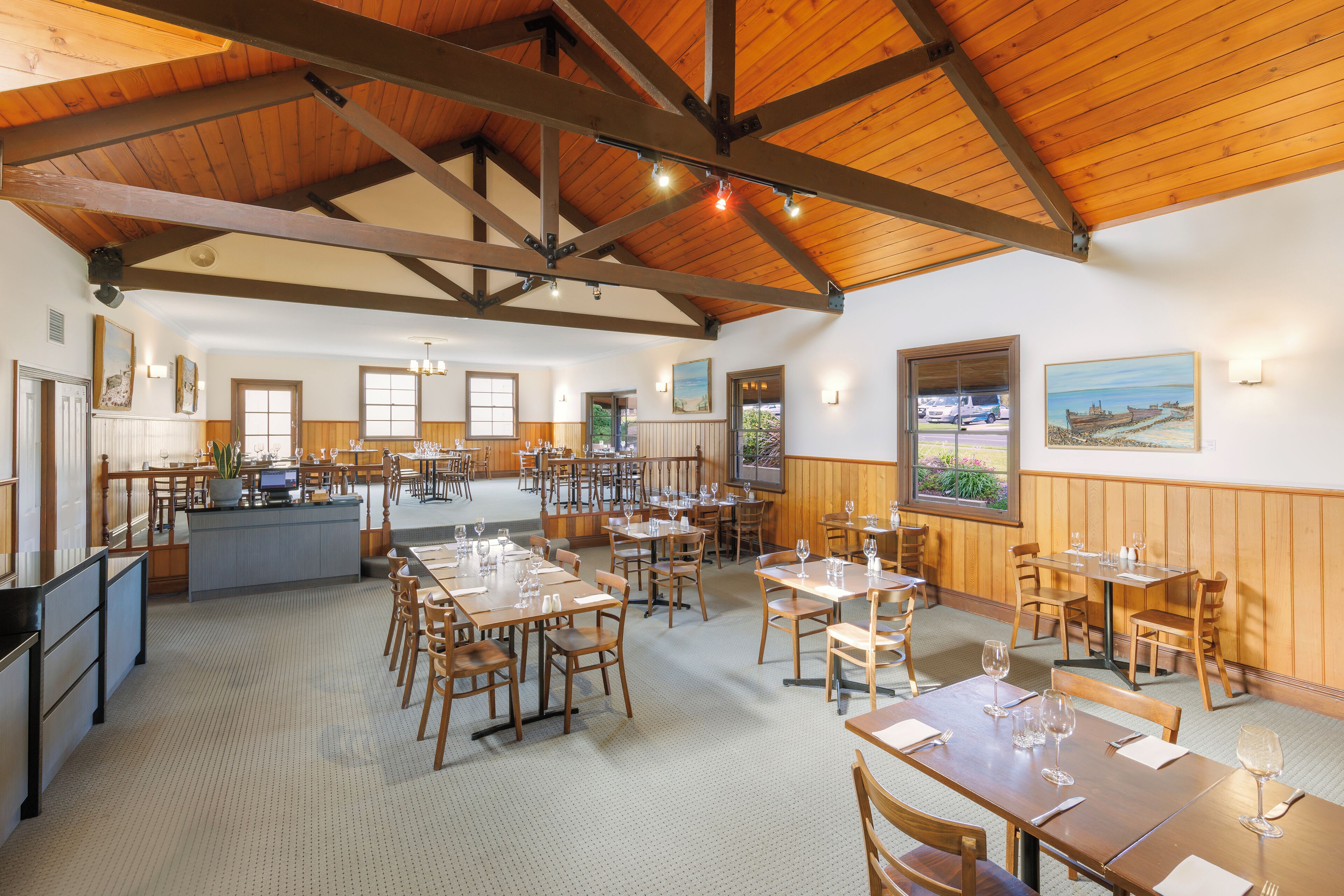 A spacious restaurant with wooden beams and a high ceiling, featuring multiple dining tables and chairs, a bar area, and large windows providing natural light.