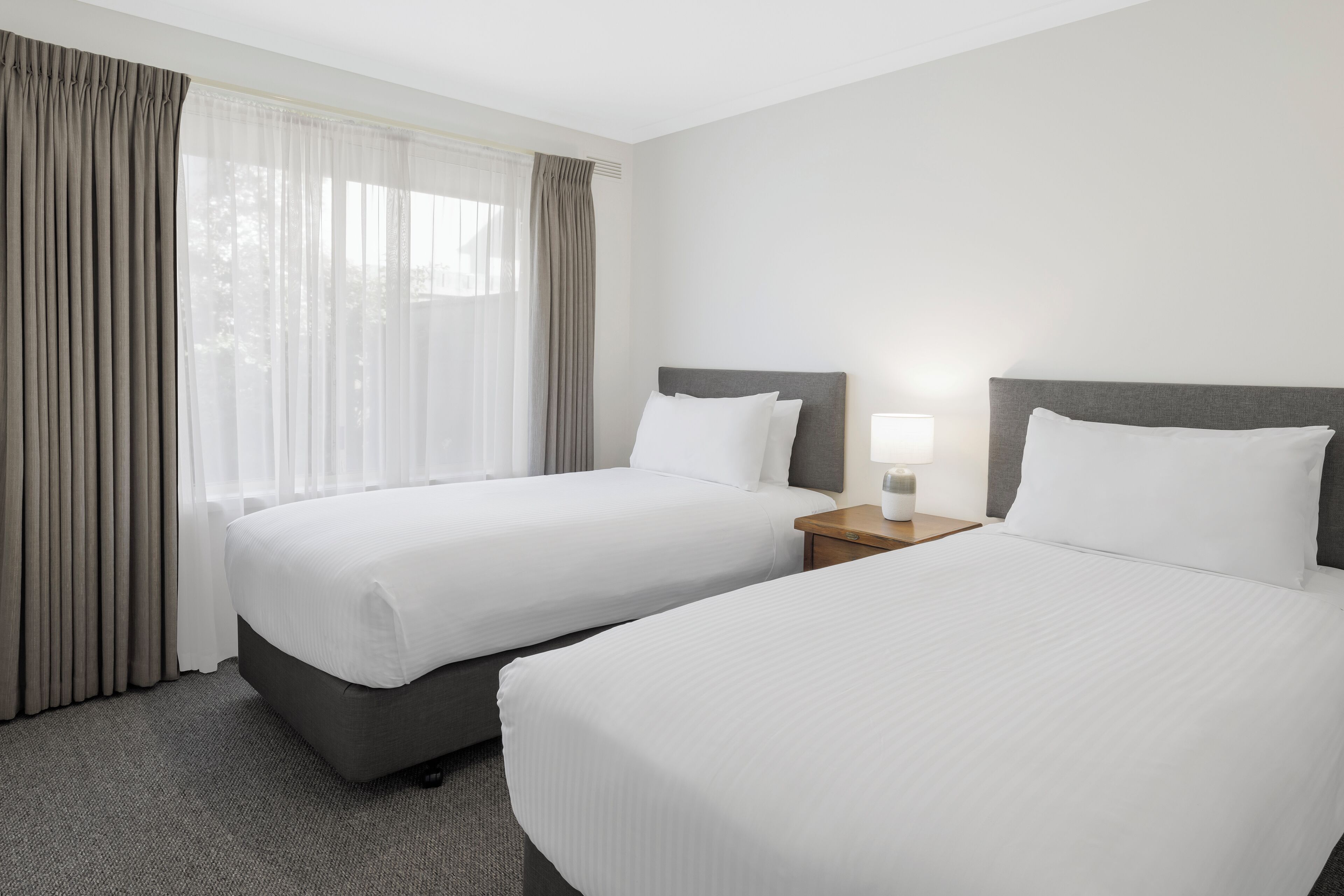 A hotel room with two beds, a nightstand with a lamp, and a window with curtains.