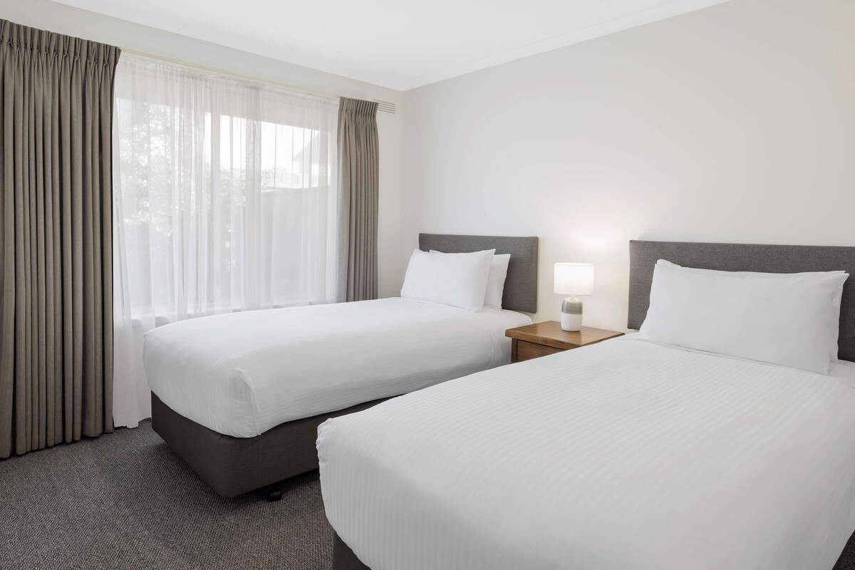 A hotel room with two beds, a nightstand with a lamp, and a window with curtains.