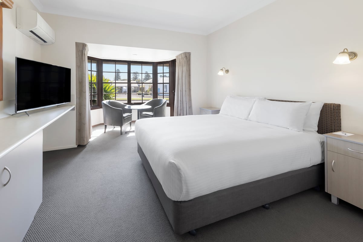 A hotel room with a large bed, a desk with a flat-screen TV, a small table with two chairs, and a window with curtains.