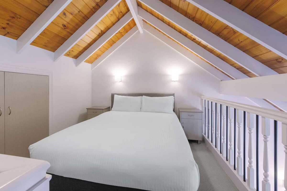 A bedroom with a sloped ceiling, a bed, bedside tables, a wardrobe, and a railing.