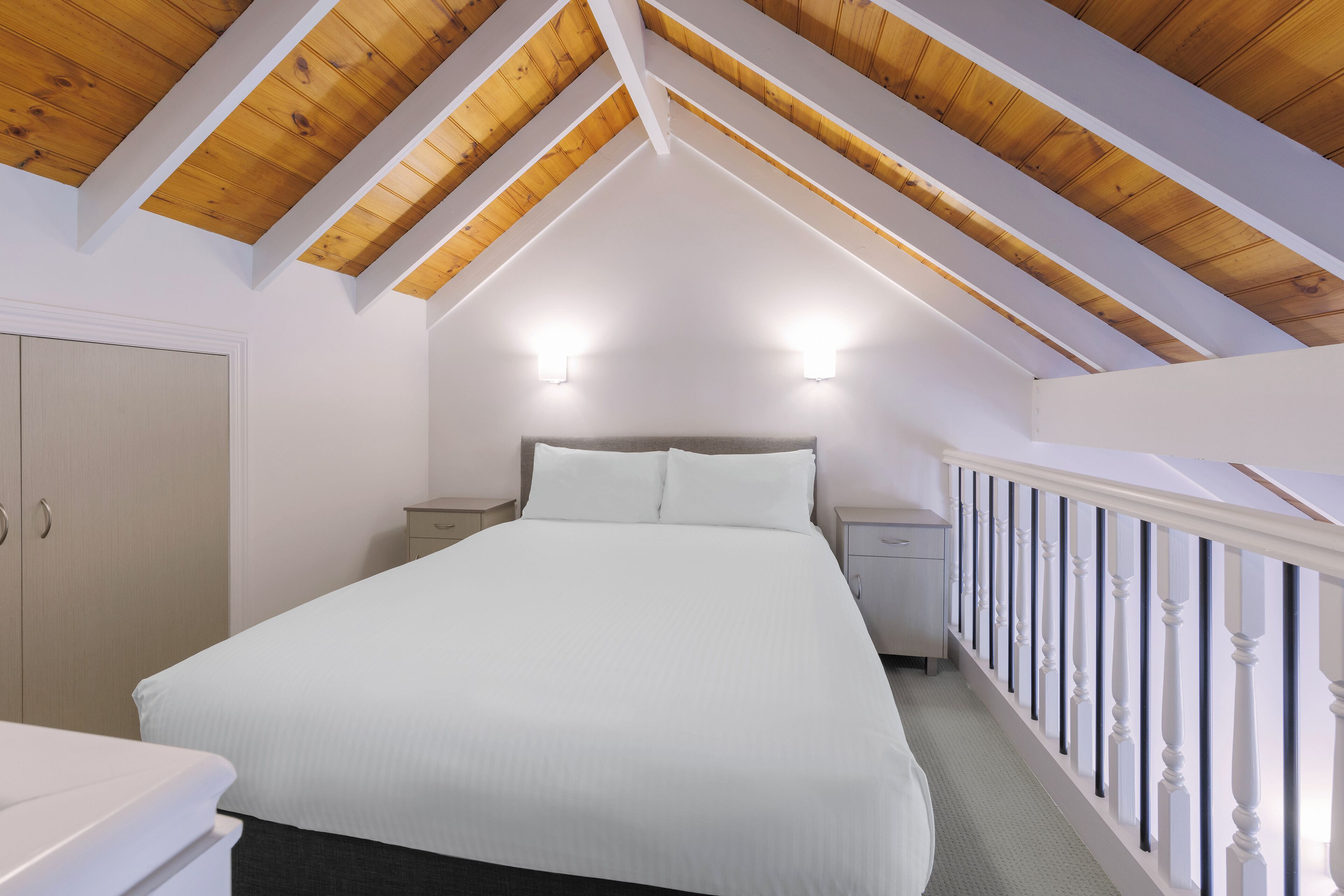 A bedroom with a sloped wooden ceiling, a bed with white bedding, two bedside tables, and a white railing along the staircase.