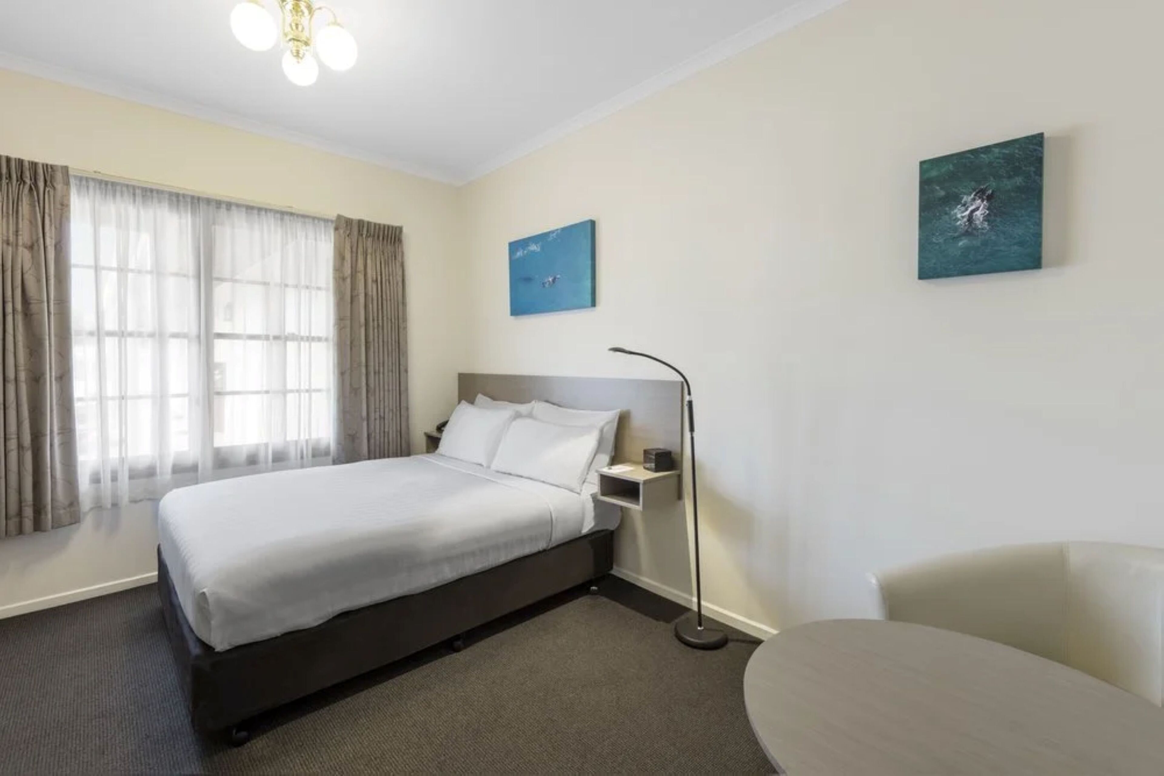 A hotel room with a bed, a bedside table, a chair, and two paintings on the wall.