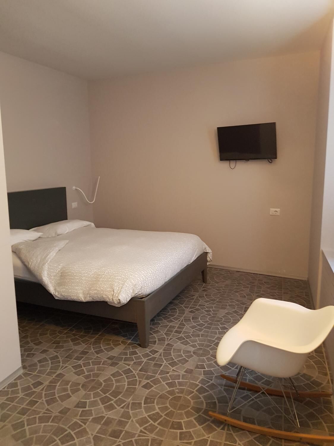 Ein kleines Zimmer mit einem Bett, einem an der Wand befestigten Fernseher und einem weißen Stuhl.
