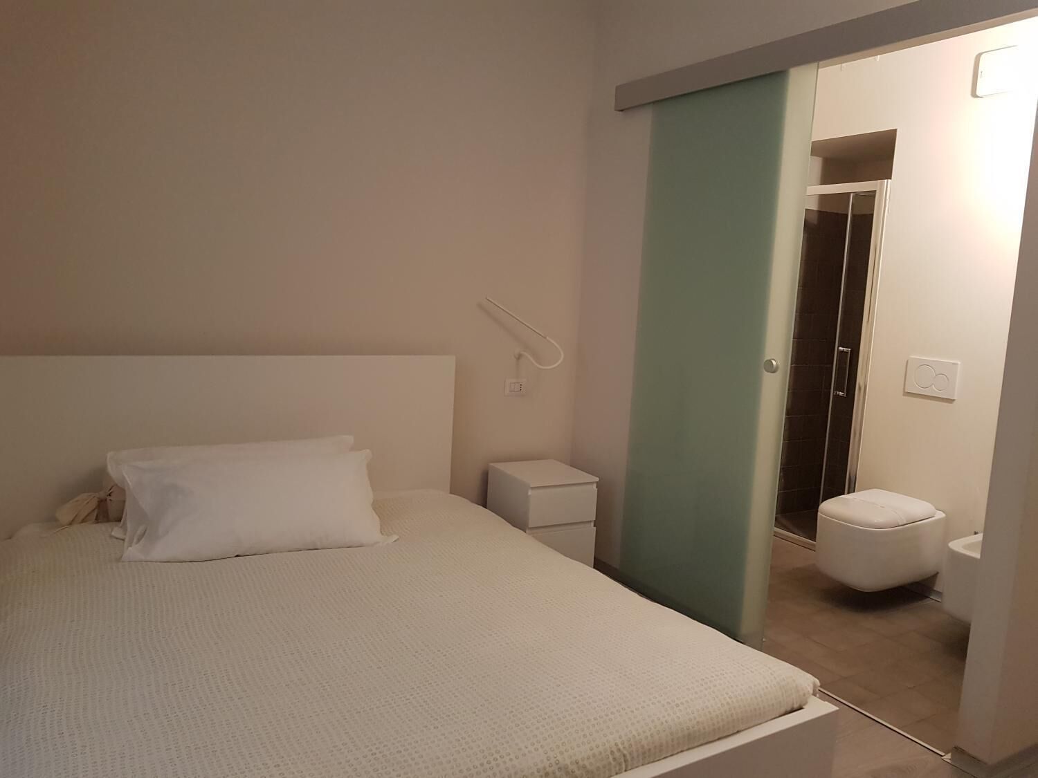 Une chambre à coucher comprenant un lit, une table de chevet, une commode, une lampe fixée au mur, des toilettes et un espace douche.