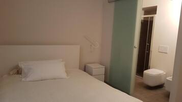 Une chambre à coucher comprenant un lit, une table de chevet, une commode, une lampe fixée au mur, des toilettes et un espace douche.