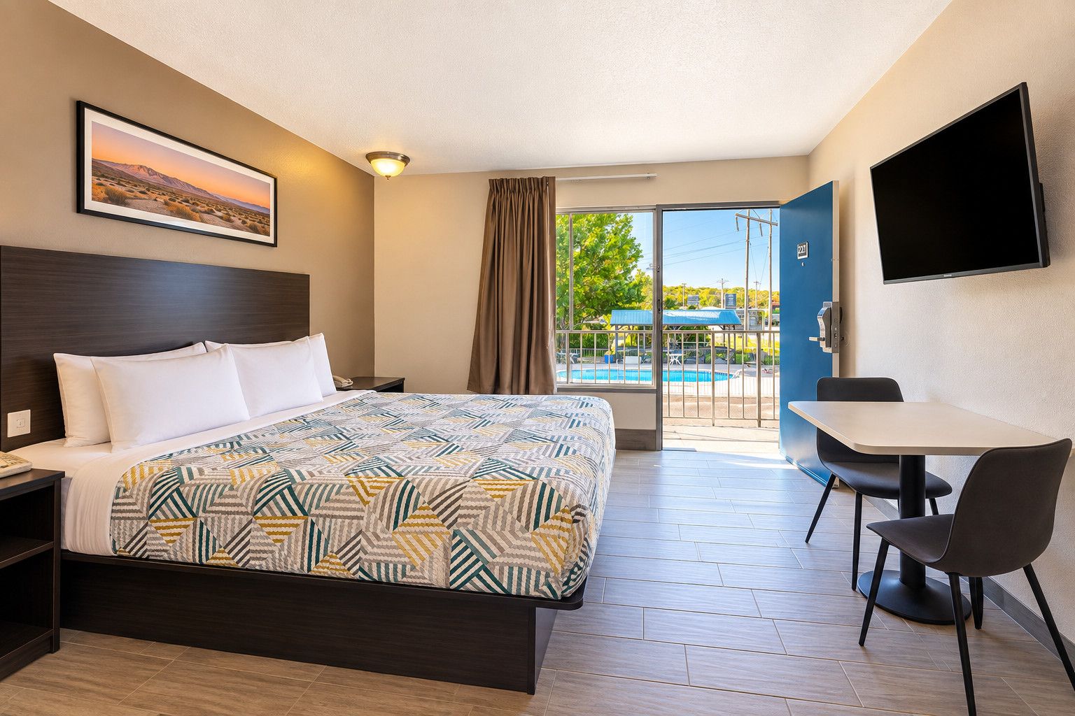 A hotel room with a bed, a desk, a chair, a TV, a window with a view of a pool, and a door.