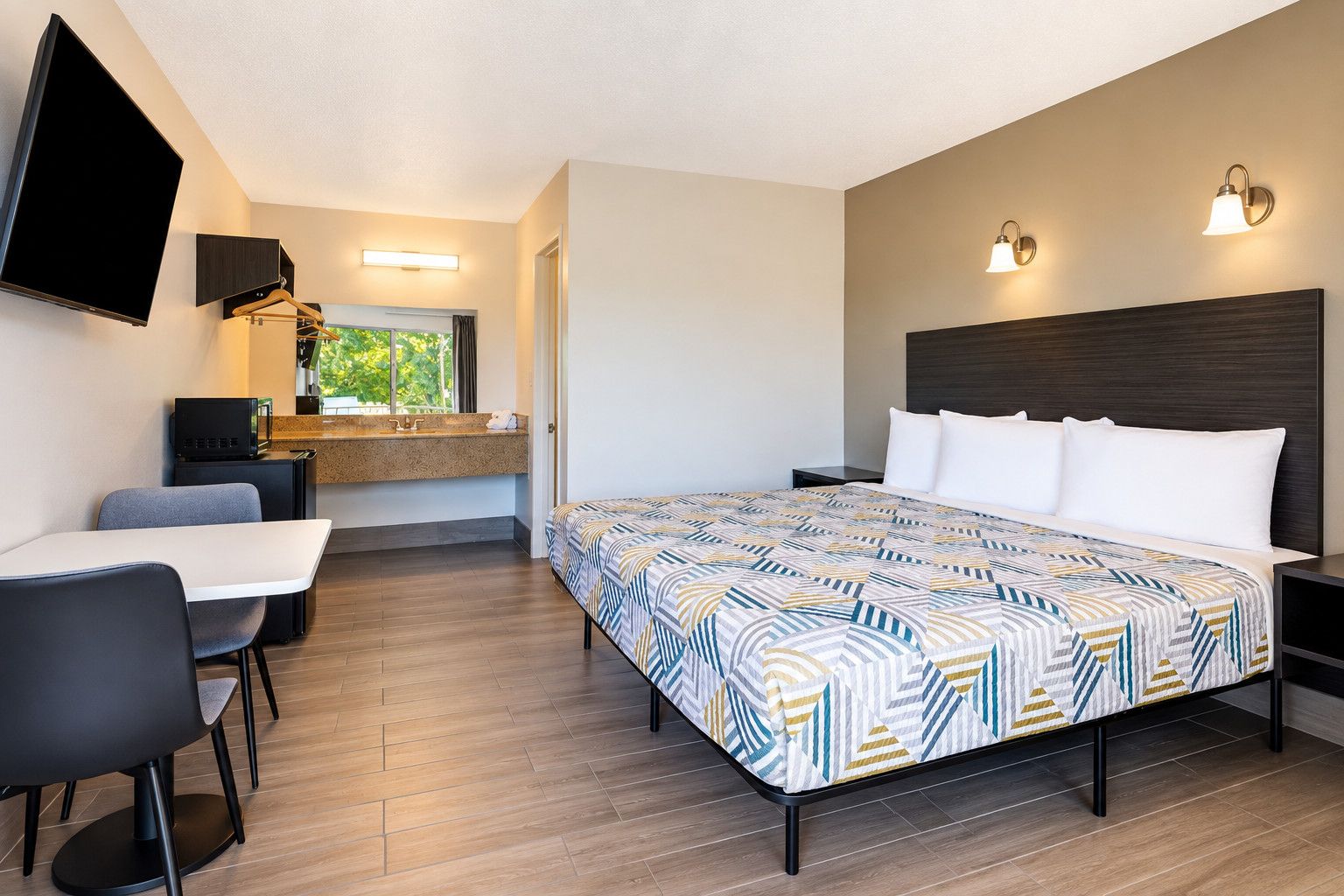 A hotel room with a bed, bedside tables, a desk with a chair, a flat-screen TV mounted on the wall, and a window with a view of greenery.