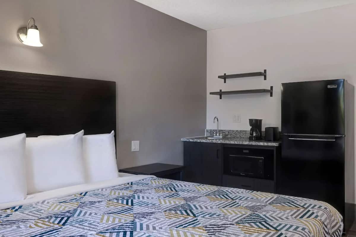 A hotel room with a bed, a nightstand, a built-in desk, a refrigerator, and a microwave.