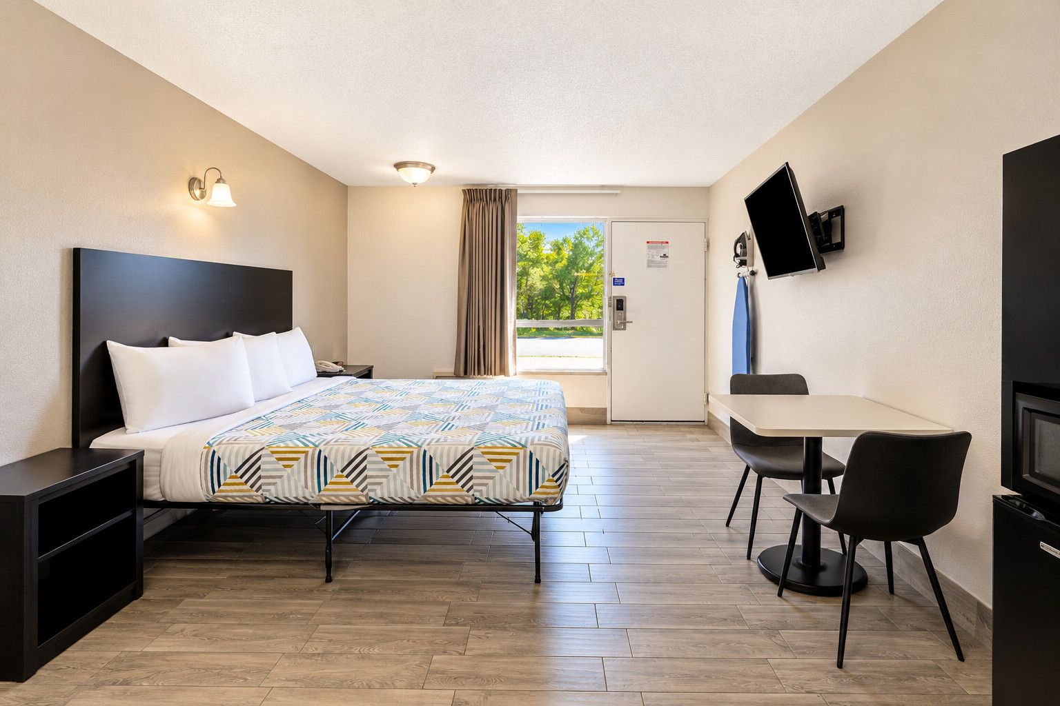 A hotel room with a bed, bedside table, desk, chair, television, and a door leading to a balcony.
