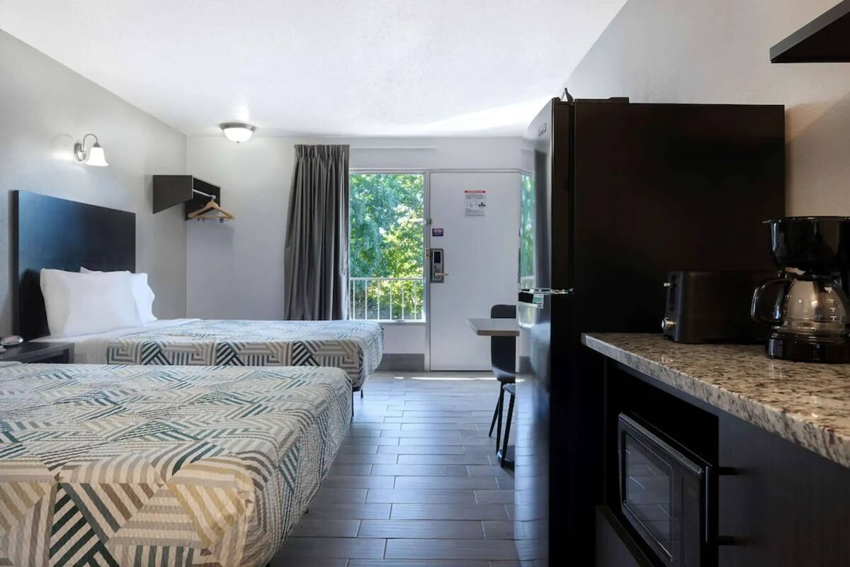 A hotel room with two beds, a kitchenette, a dining area, and a sliding glass door to the outside.