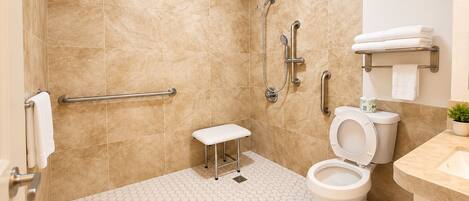 Un baño con inodoro blanco, ducha con barras de apoyo y un pequeño banco blanco.