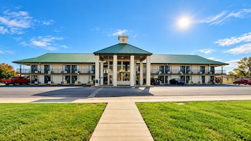 A two-story building with a central entrance, a green roof, and a parking lot with cars.